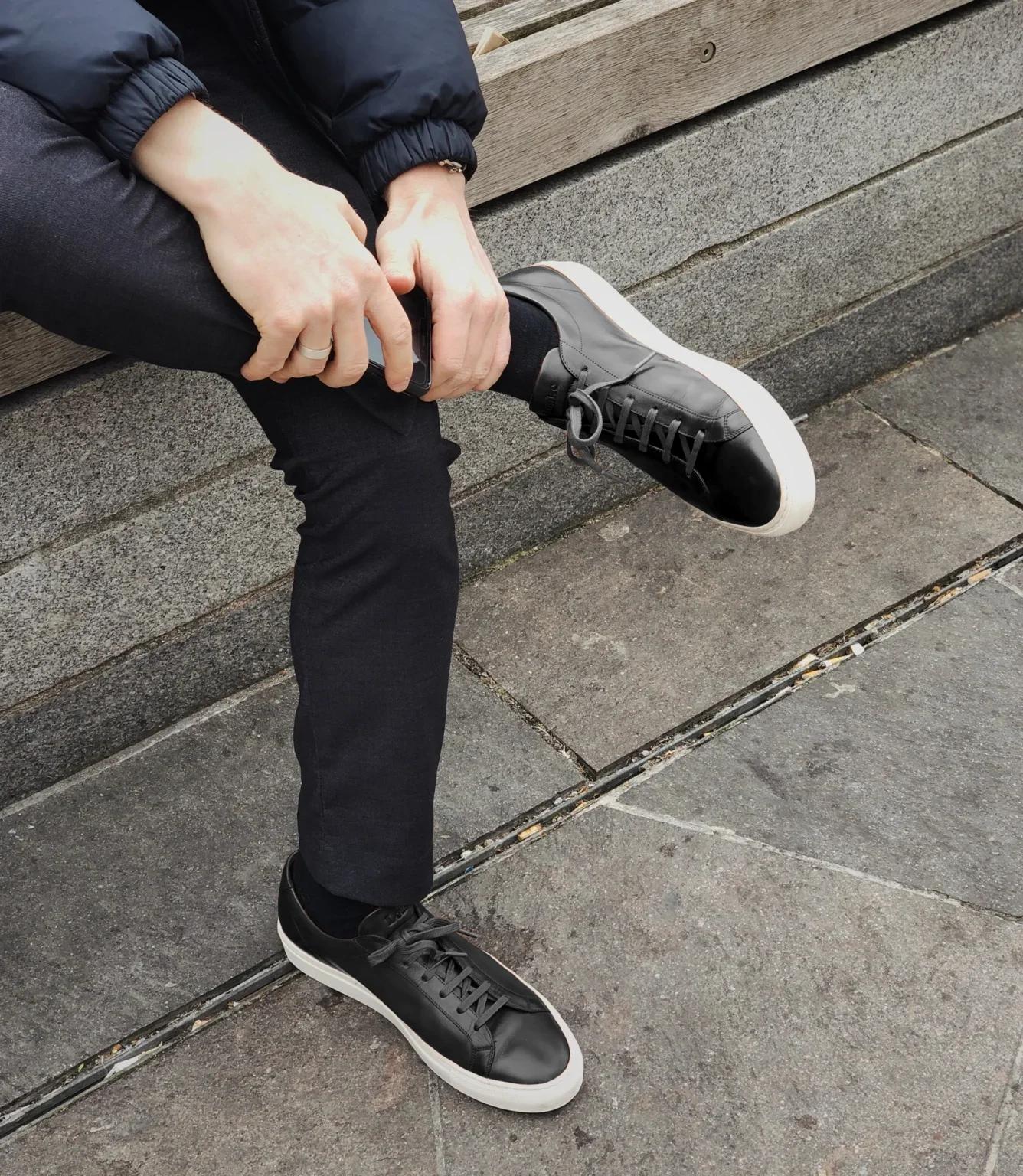 A man sitting down wearing navy blue chino trousers wearing his Loake Sprint Trainers in Black Leather.