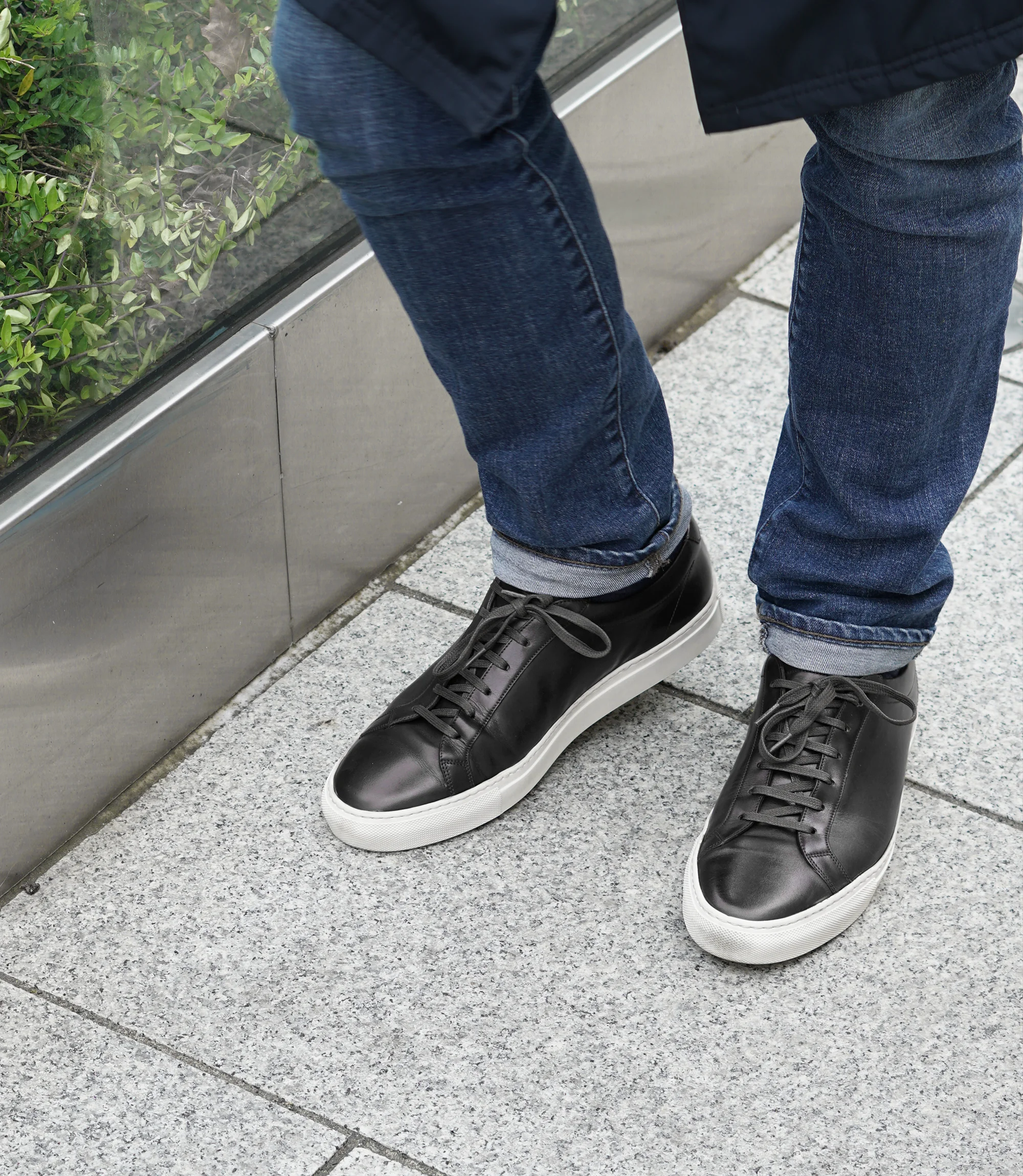 A man wearing navy denim trousers standing walking down a grey tiled floor, wearing his Loake Sprint Trainers in Black Leather.