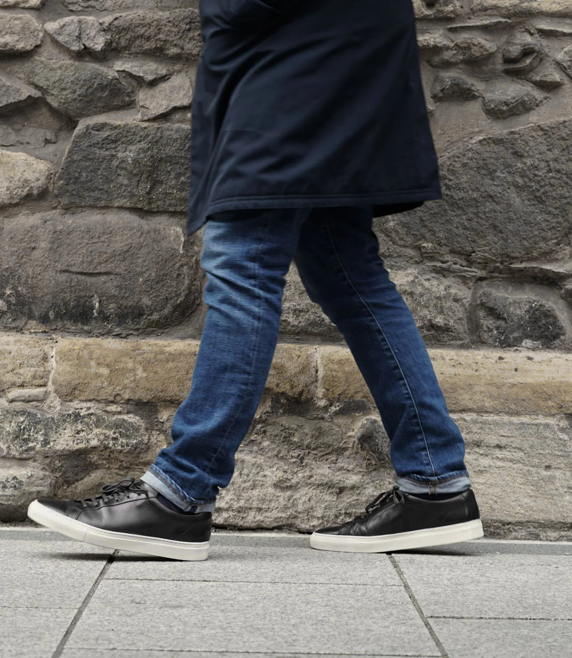 A man wearing navy denim trousers standing walking down a grey tiled floor, wearing his Loake Sprint Trainers in Black Leather.