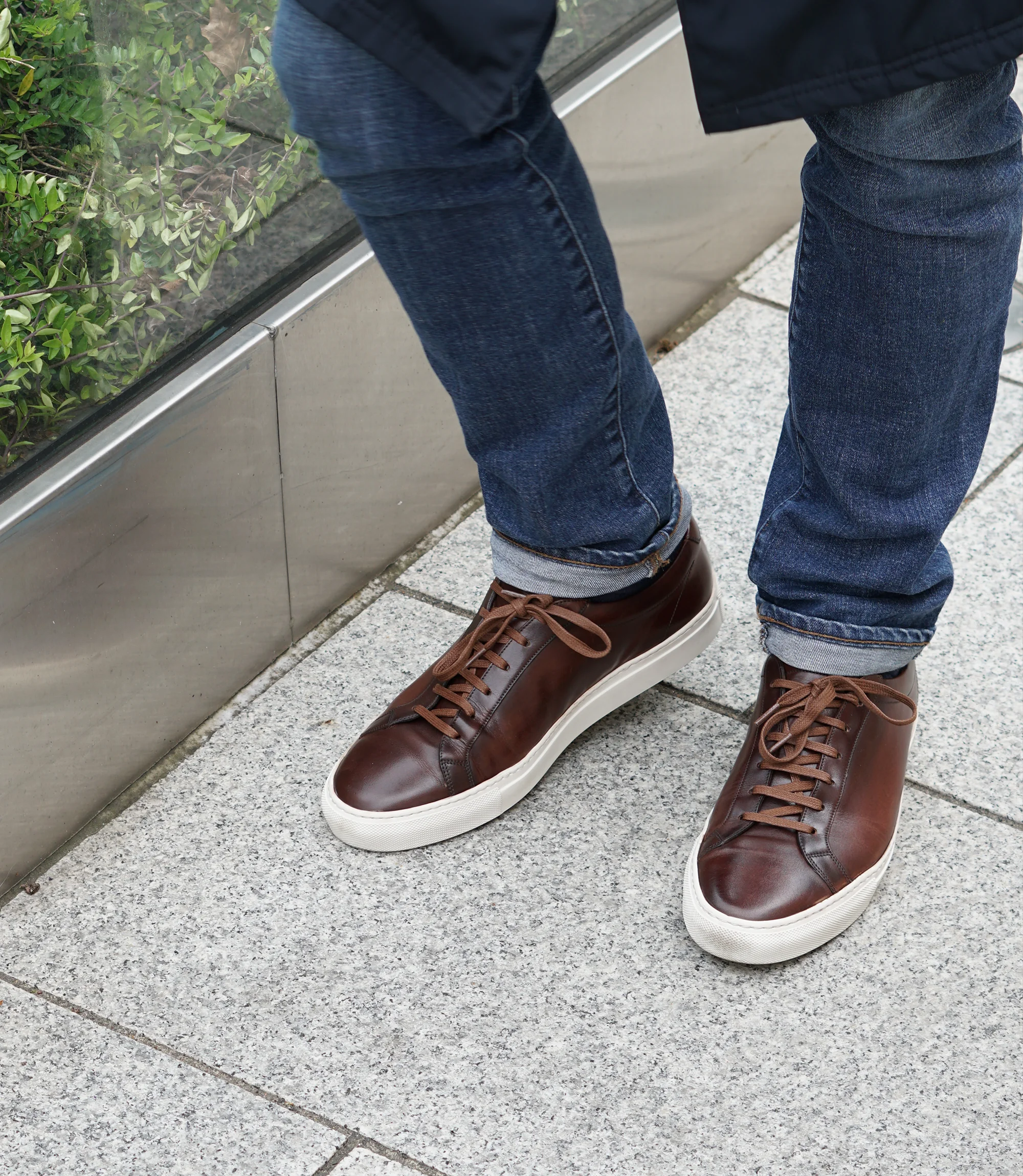 A man wearing navy denim trousers standing walking down a grey tiled floor, wearing his Loake Sprint Trainers in Dark Brown Leather.