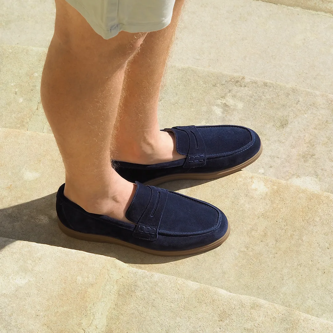 Man standing wearing navy suede Loake loafers on a sun drenched terrace, in nylon grey shorts. Shoes shown are Lucca in navy suede leather