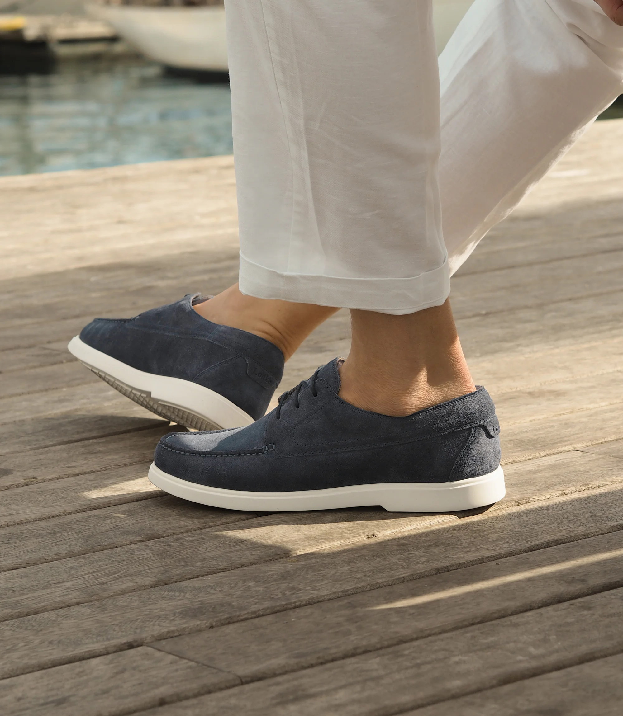 A man in white trousers, sitting down legs crossed with the sea in front of him wearing his Loake Sardinia Boat shoes in Light Blue Suede.