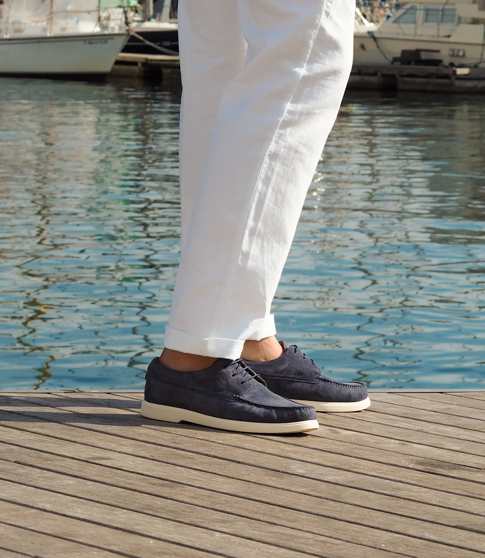 A man in white trousers, standing on a wooden floor with the sea behind him wearing his Loake Sardinia Boat shoes in Light Blue Suede.