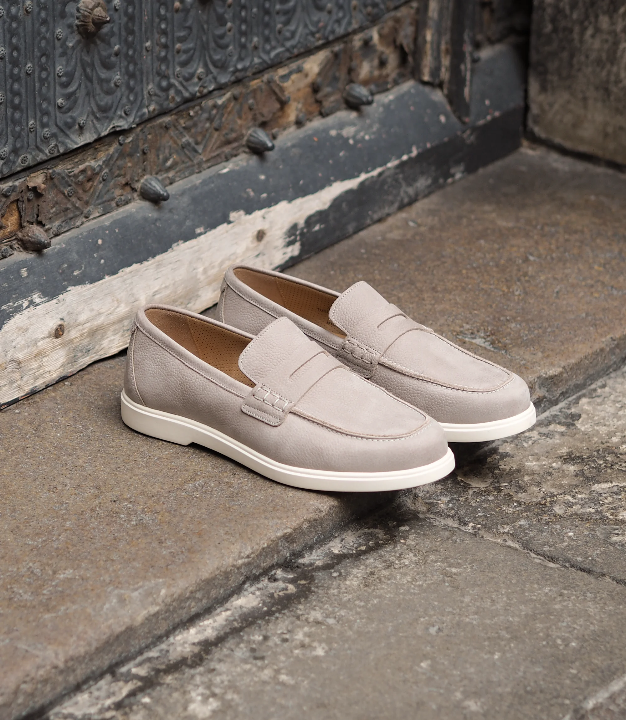 Loake Como Loafers in Grey Nubuck placed together on a concrete floor in front of a blue door.