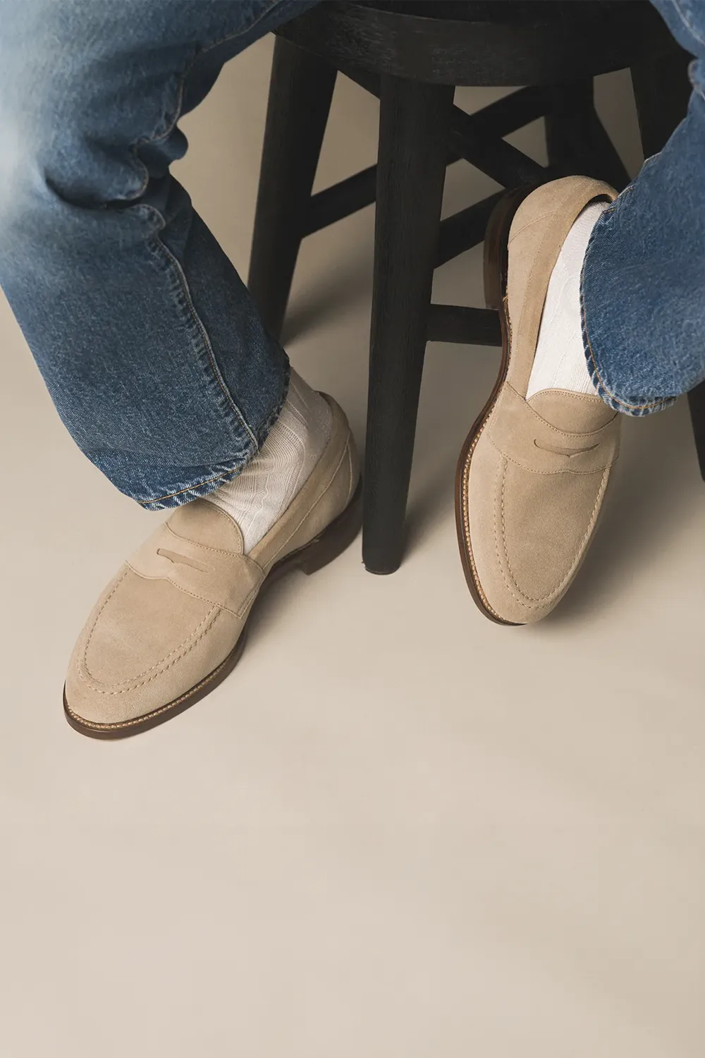 Man sat on a dark wooden stool with navy jeans on with white long socks wearing Loake Imperial sand suede loafers.