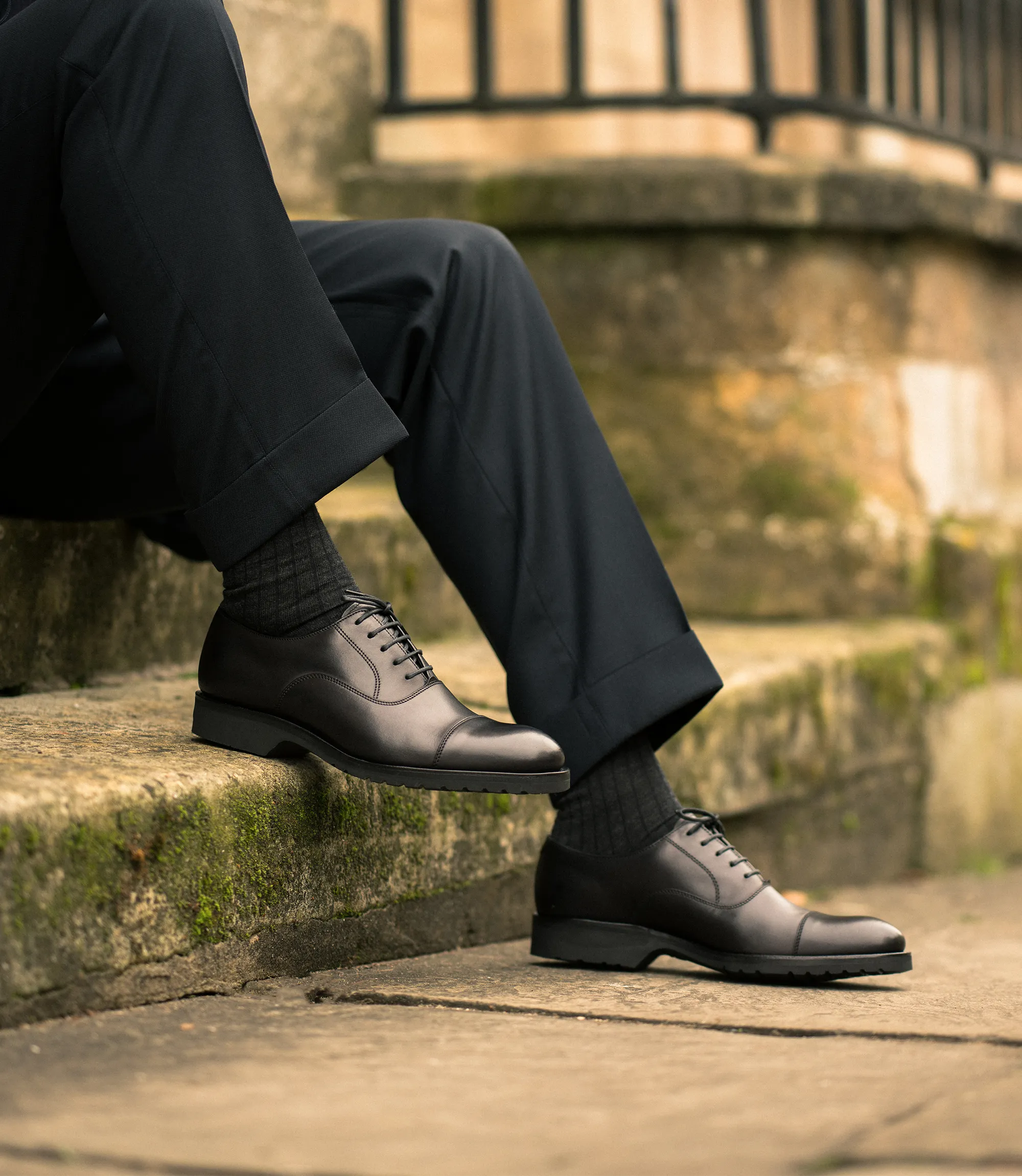 A man in navy suit trousers and jacket sitting on a weathered concrete step in a city scene, whilst wearing his Loake Hitchcock Black Leather Toe Cap Oxford Shoes.