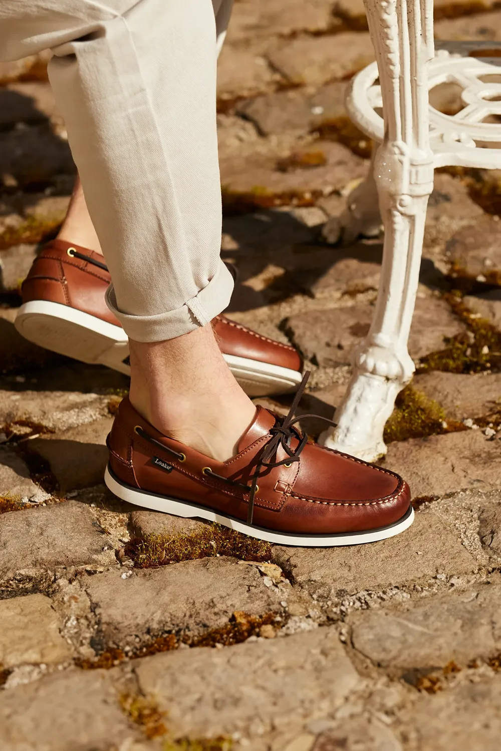 Man wearing cedar Boat Shoes, sitting at white cafe table on a cobbled street. Shoes shown are Loake 528 in Cedar leather
