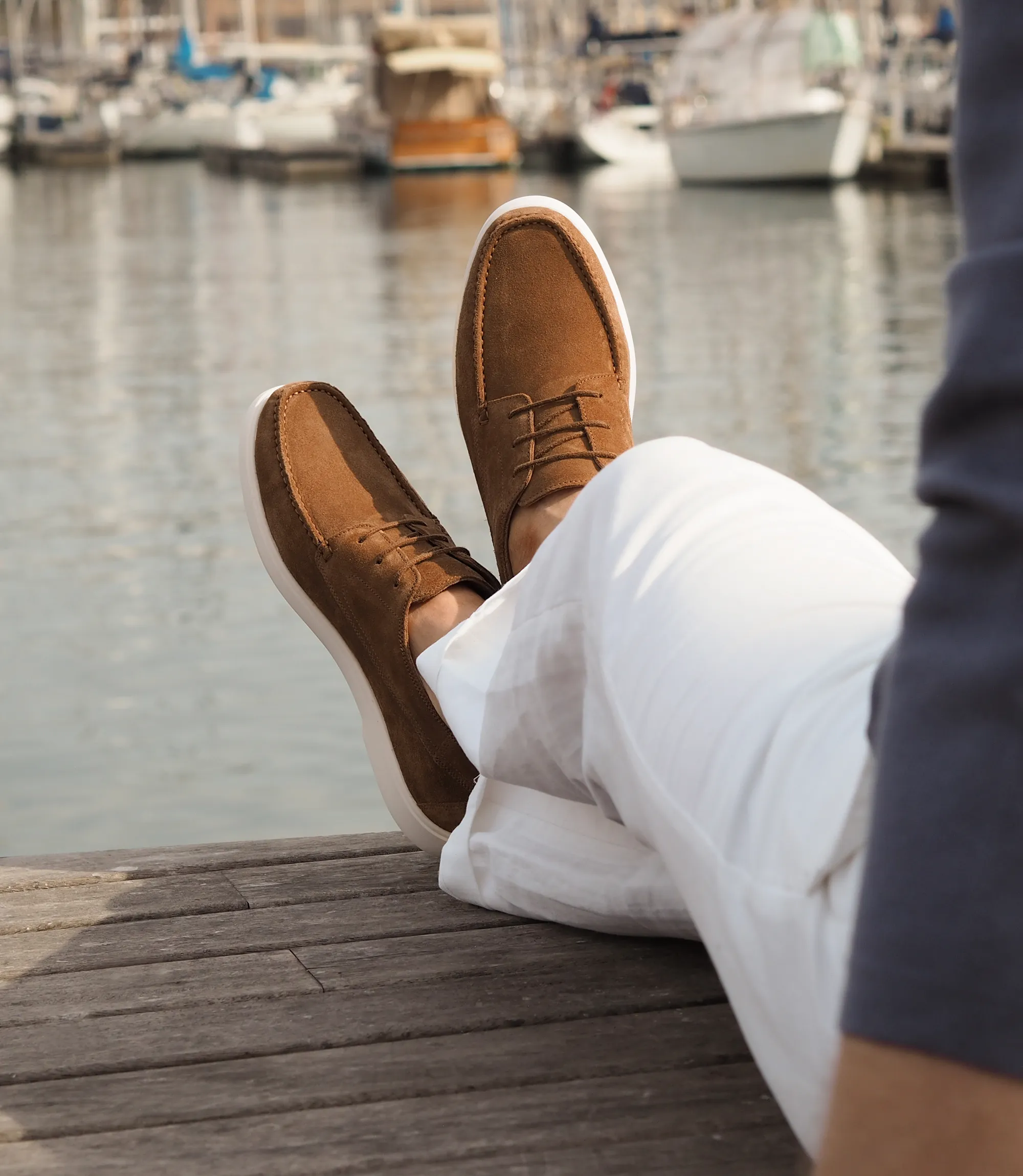Man wearing white linen trousers and navy sweatshirt laying on a wooden deck by the sea, with his legs crossed. He is wearing a pair of Loake Sardinia Boat Shoes in Chestnut Brown Suede