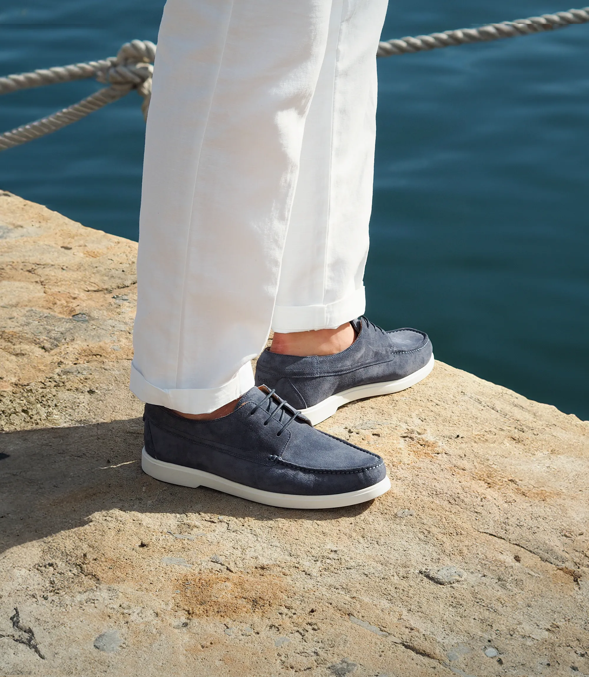 Man standing on a rock by the sea, with the sunset creating a silhouette of himself and his Loake Sardinia Boat Shoes in Light Blue Suede