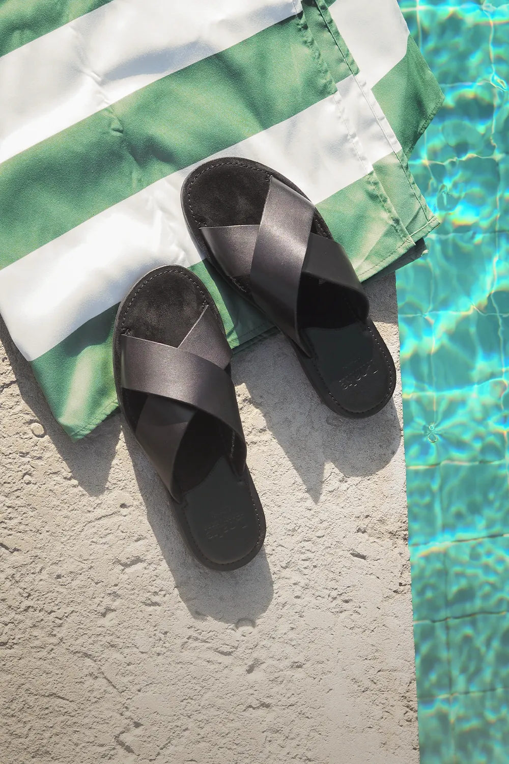 Pair of black Loake Gozo sandals sat on a green and white striped beach towel on the side of a swimming pool