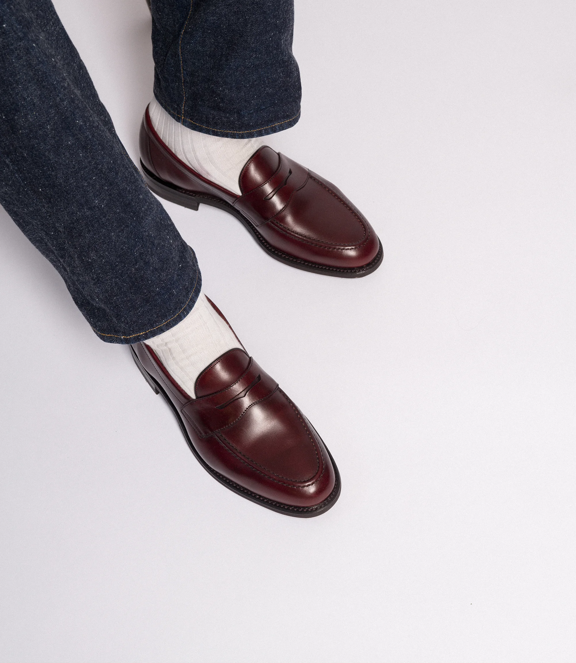 A man in navy blue denim trousers, standing on a grey backdrop floor wearing his Loake Burgundy Leather Imperial Loafers.