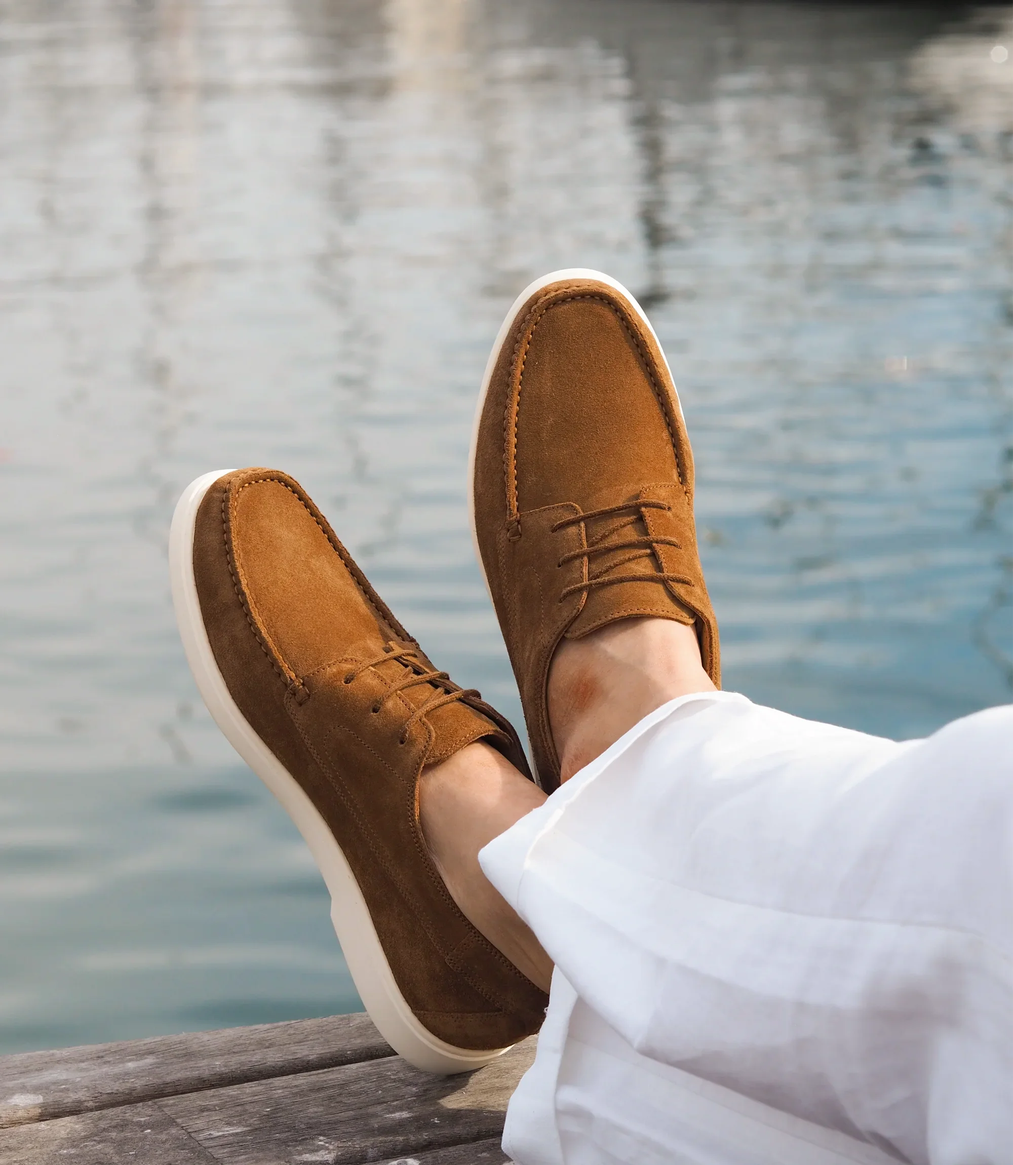 A man in white trousers, sitting down legs crossed with the sea in front of him wearing his Loake Sardinia Boat shoes in Chestnut Suede.