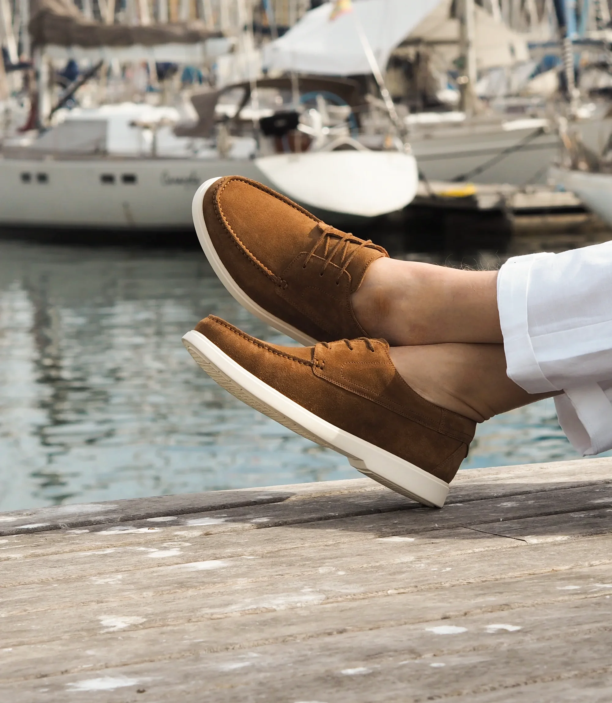 A man in white trousers, sitting down legs crossed with the sea in front of him wearing his Loake Sardinia Boat shoes in Chestnut Suede.