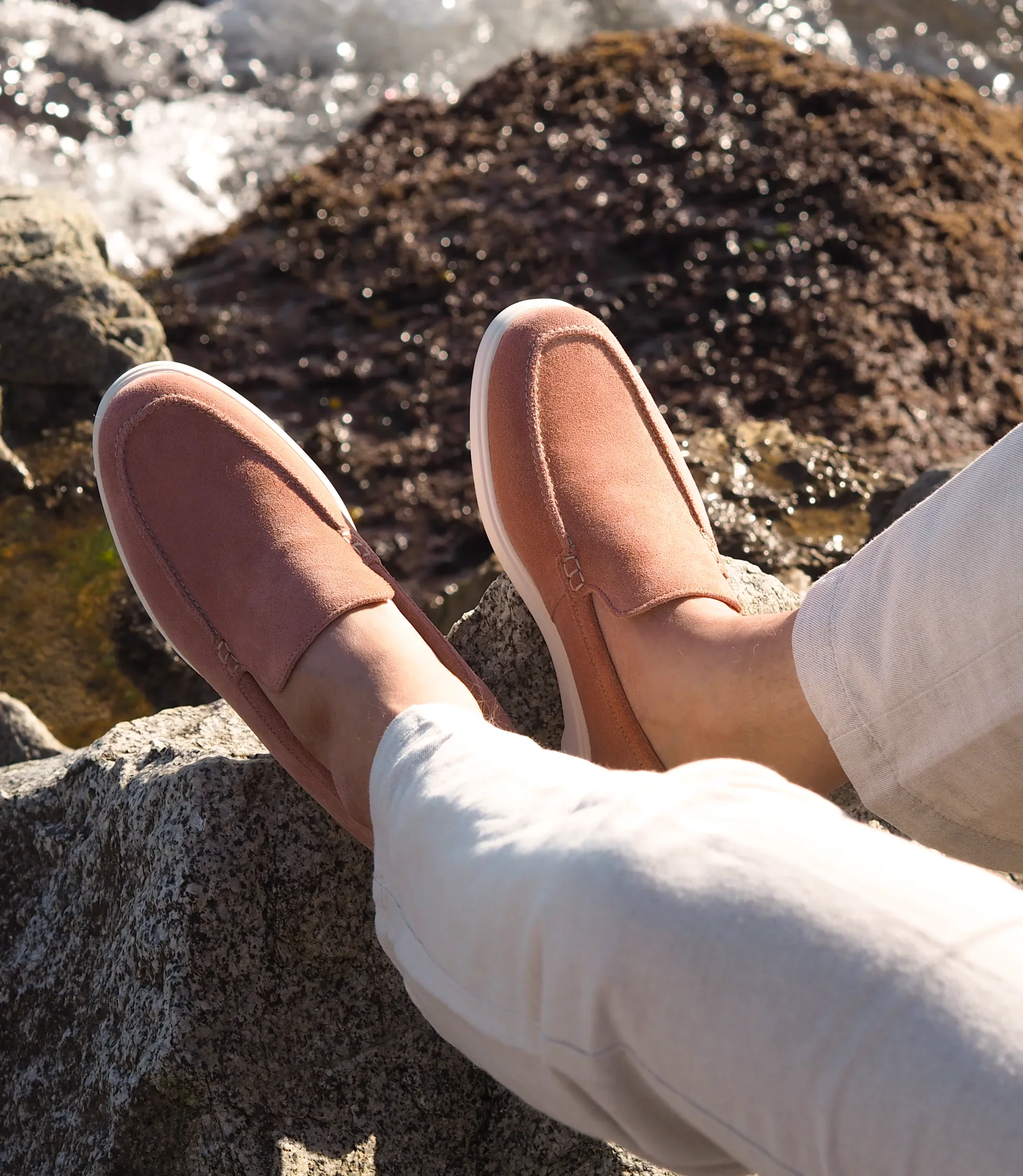 A man wearing white denim trousers sat overlooking the sea on rocks wearing his Loake Tuscany Loafers in Pink Suede.