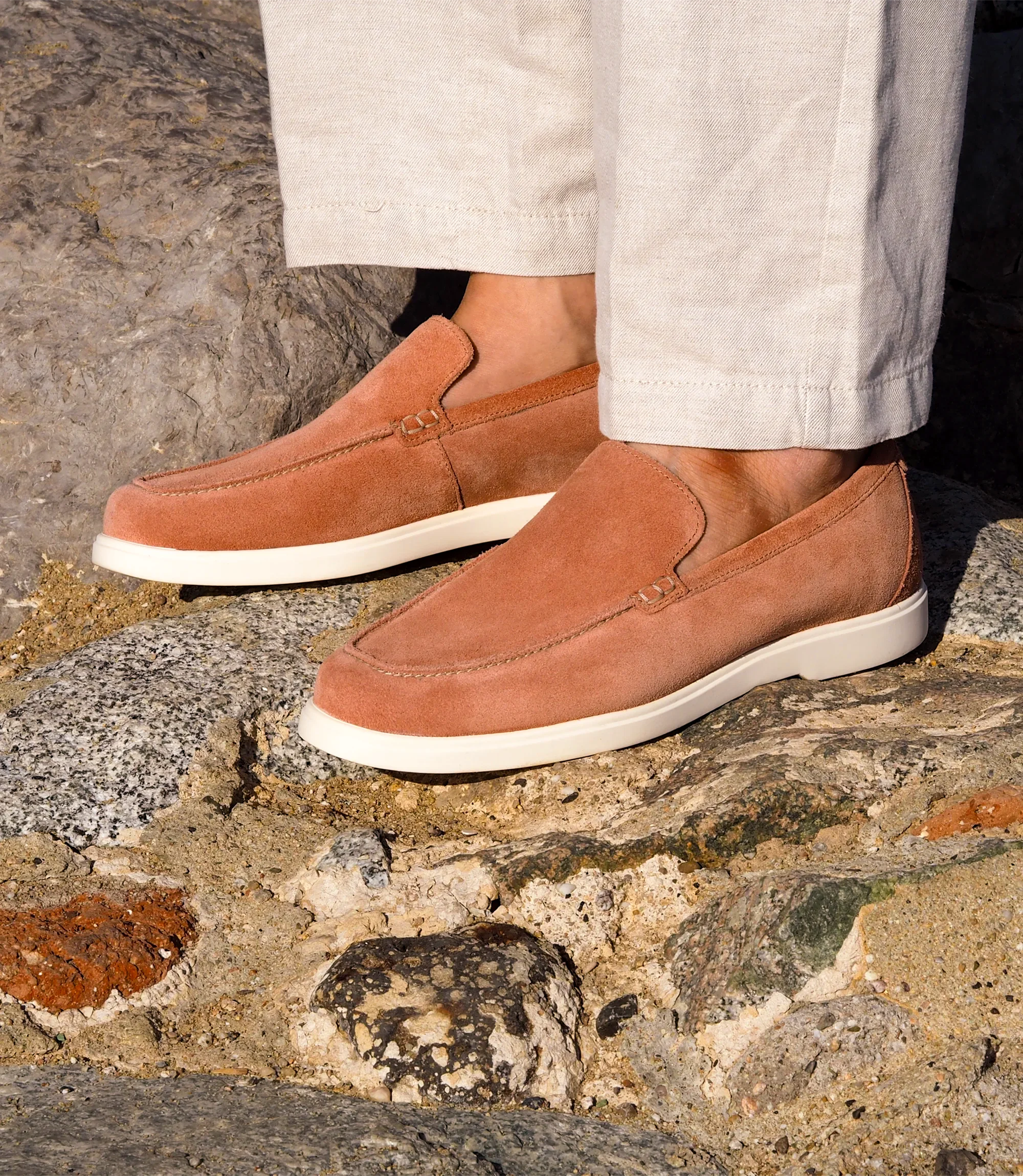 A man wearing white denim trousers standing overlooking the sea on rocks wearing his Loake Tuscany Loafers in Pink Suede.