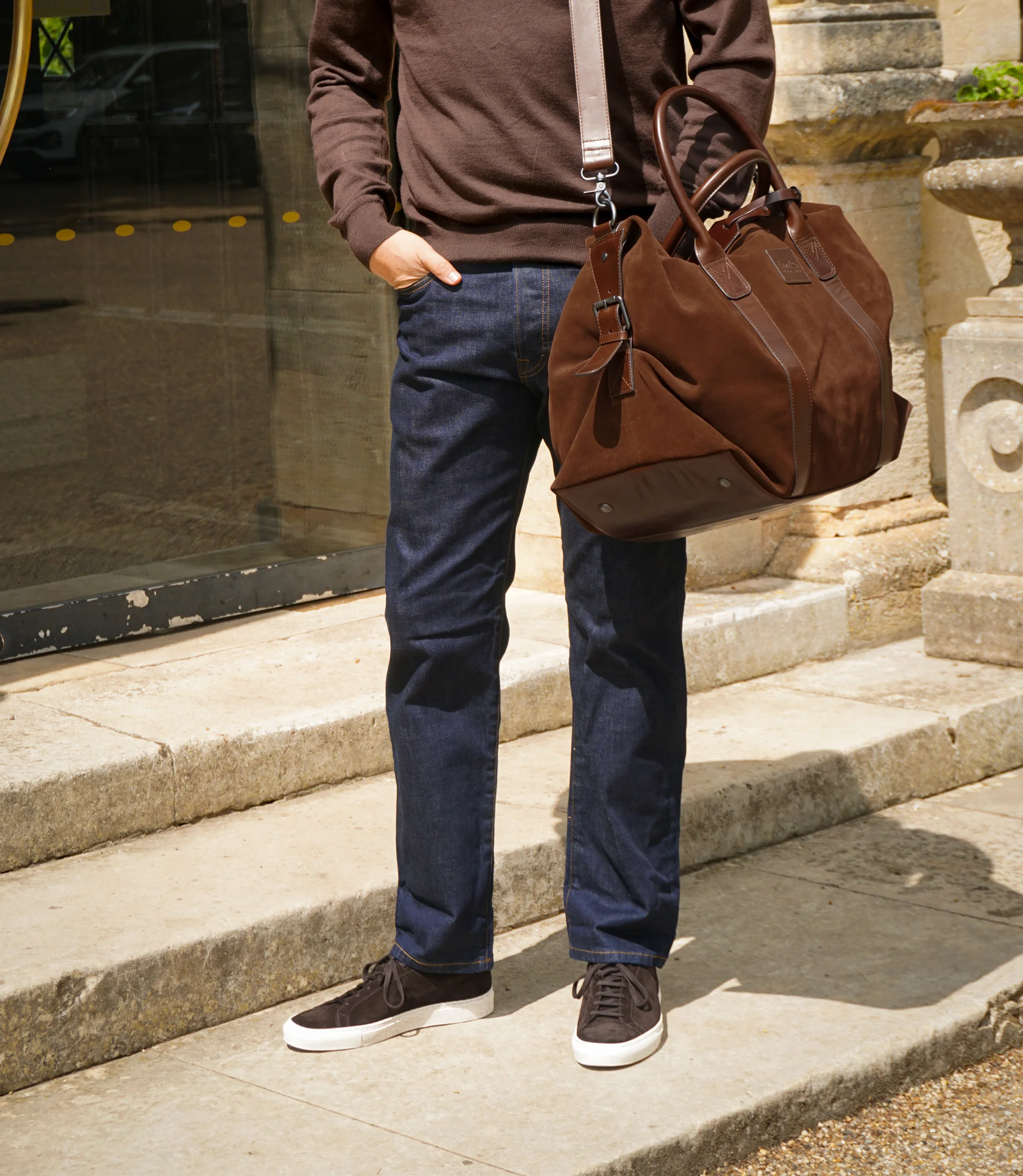 A man wearing a brown sweatshirt and navy jeans standing outside on concrete steps holding his Loake Balmoral weekend bag in Polo Suede.