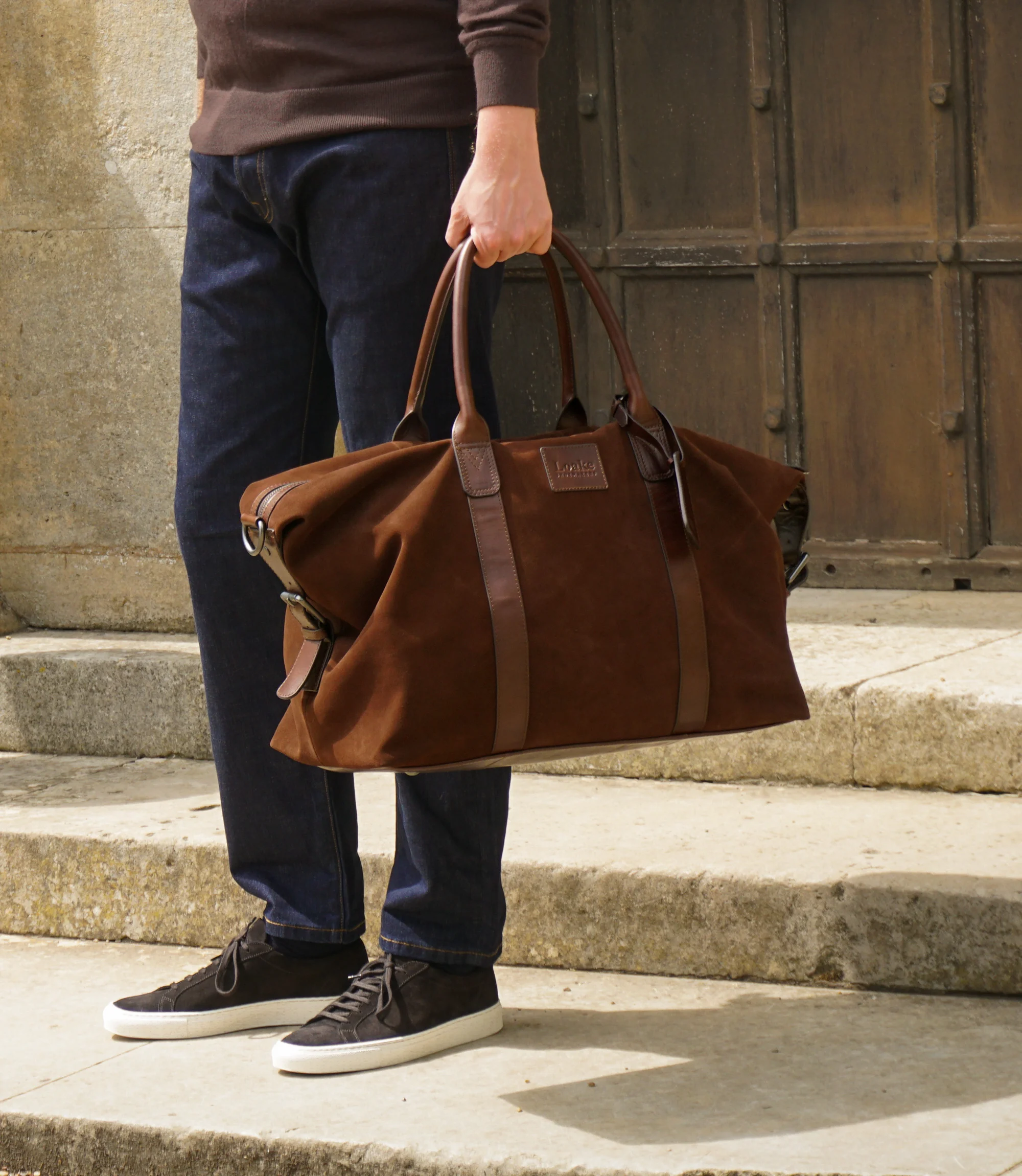 A man wearing a brown sweatshirt and navy jeans standing outside on concrete steps holding his Loake Balmoral weekend bag in Polo Suede.