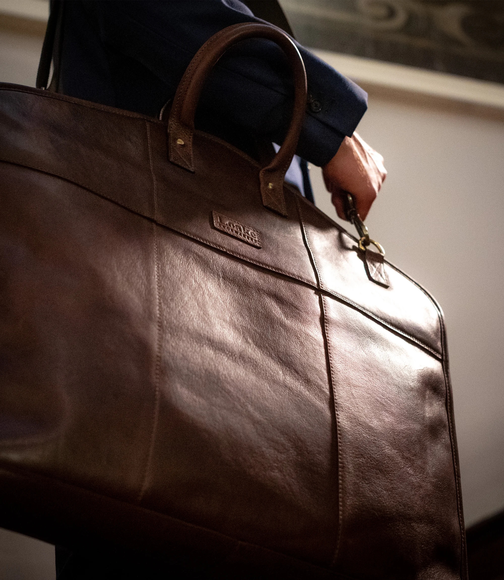 A man in navy suit walking up stairs holing his Loake London Suit Carrier in Brown Leather.