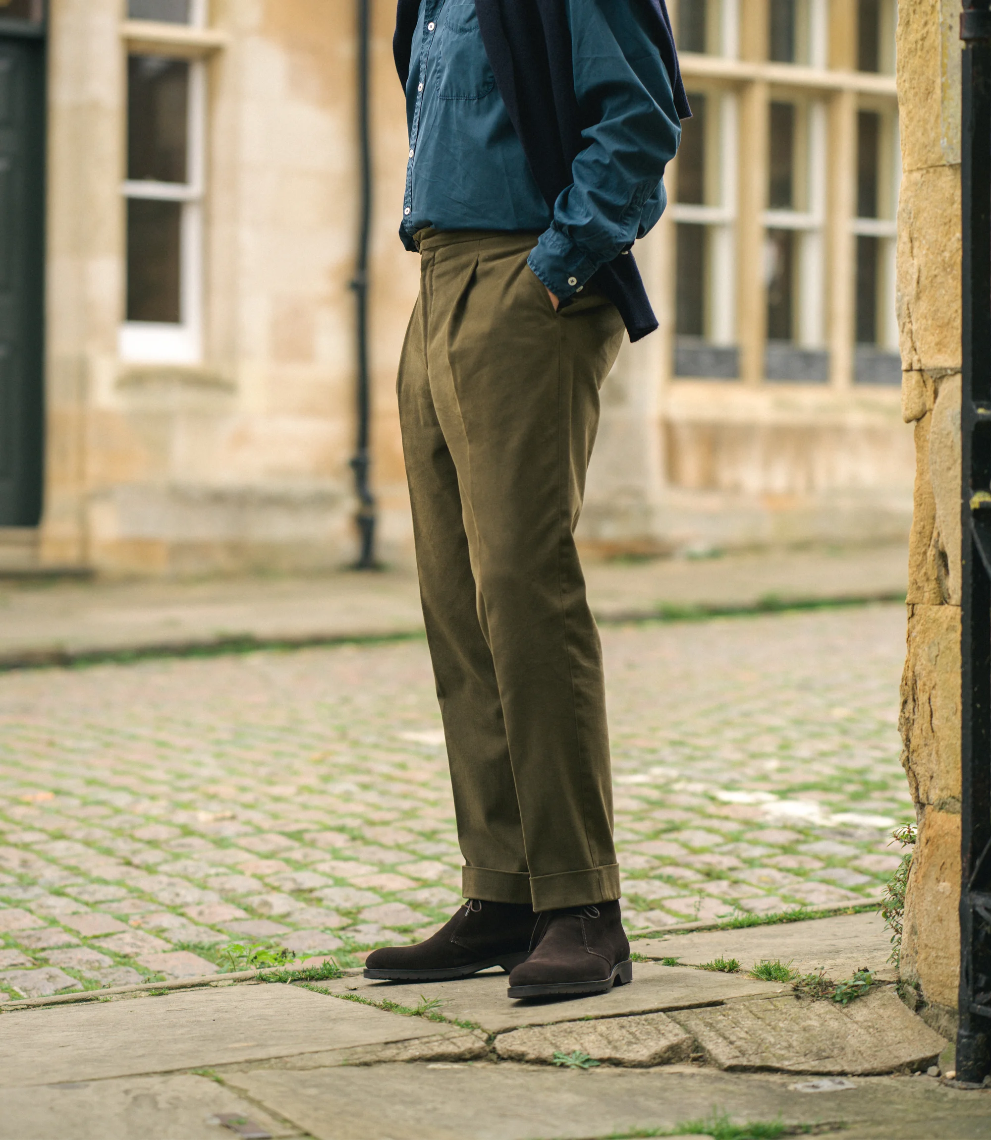 A man wearing green trousers and a blue shirt standing on a cobbled street wearing his Loake Boots Altman in Dark Brown Suede.