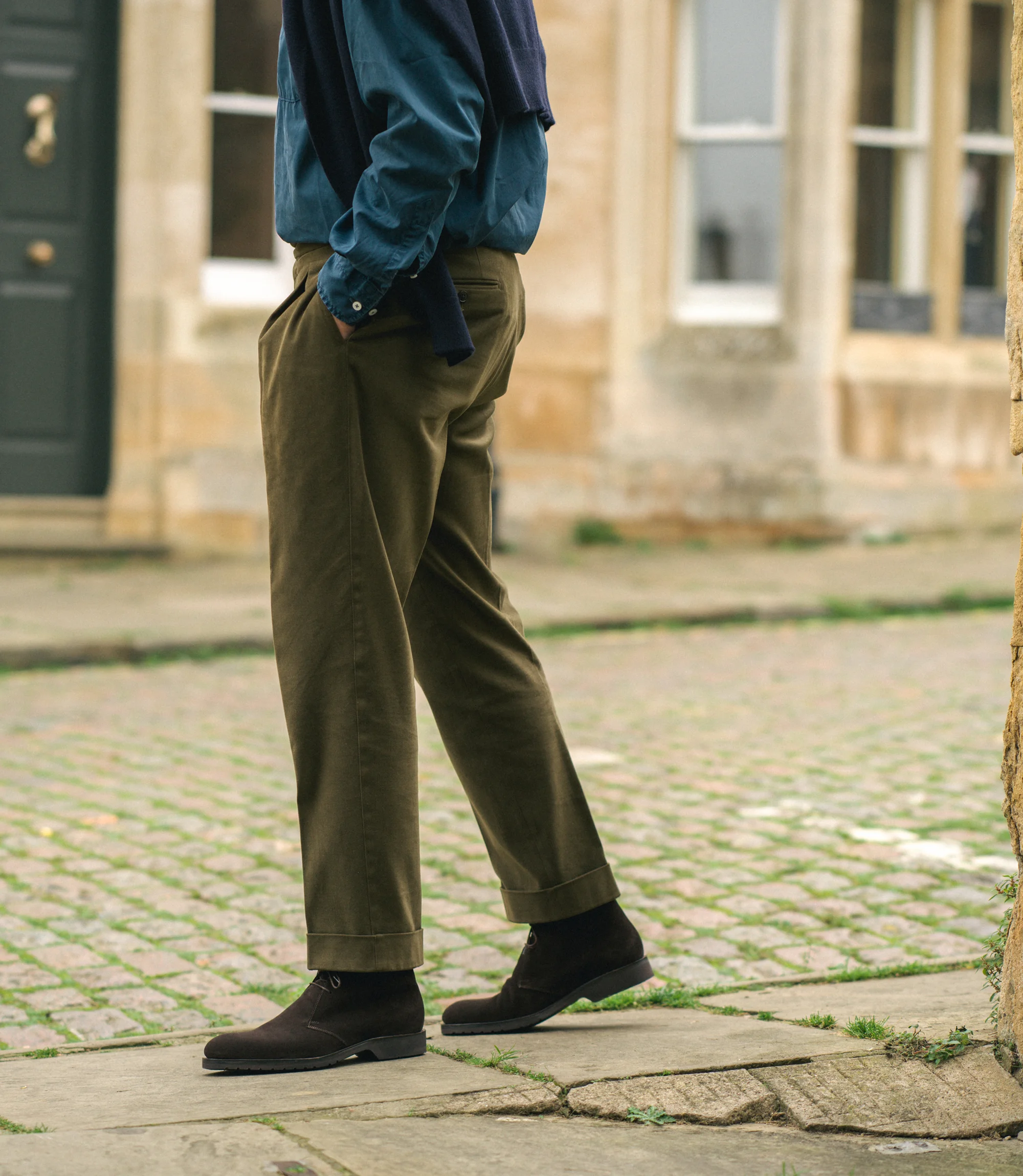 A man wearing green trousers and a blue shirt standing on a cobbled street wearing his Loake Boots Altman in Dark Brown Suede.