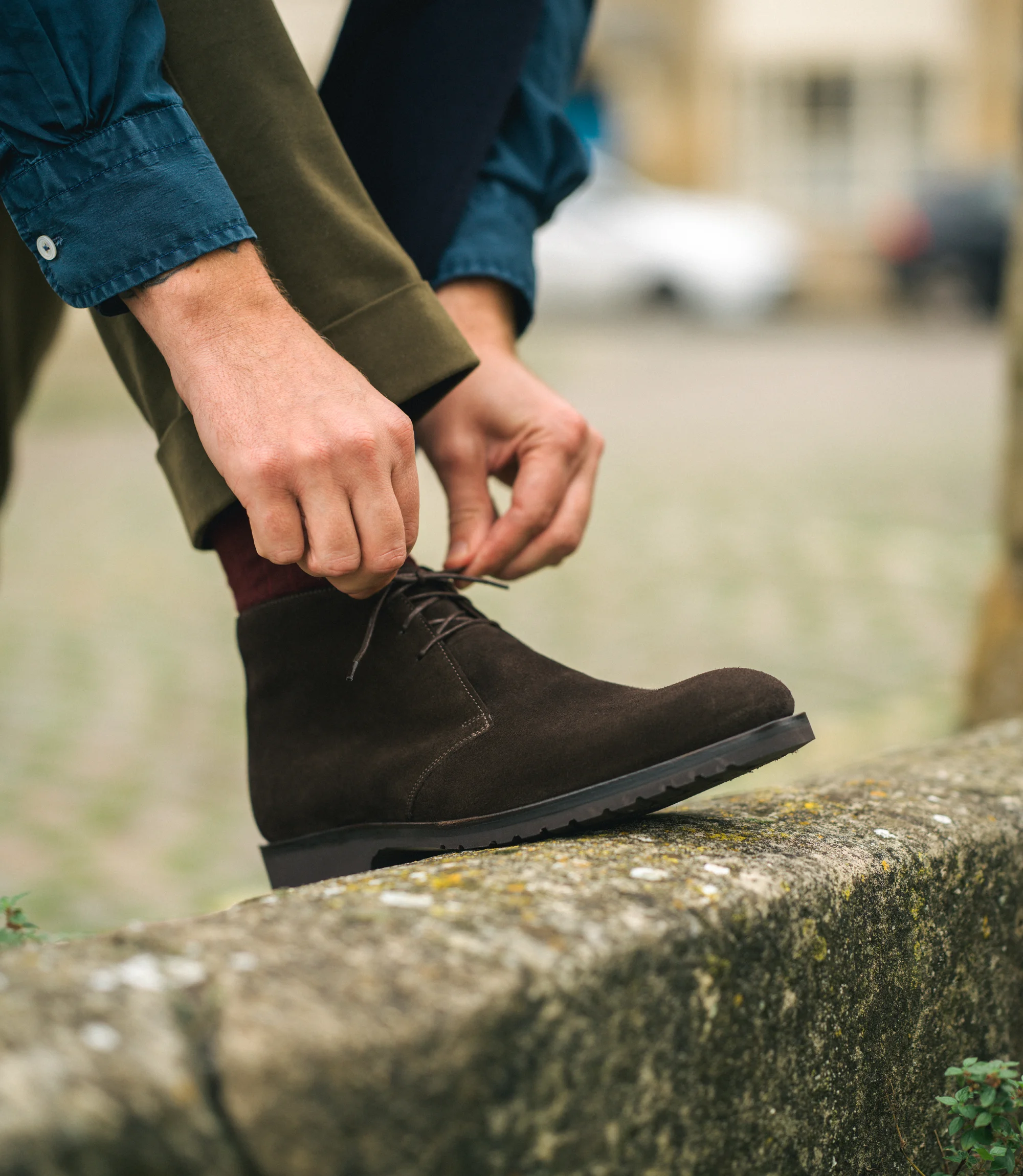 A man wearing green trousers and a blue shirt tying his laces of his Loake Boots Altman in Dark Brown Suede.