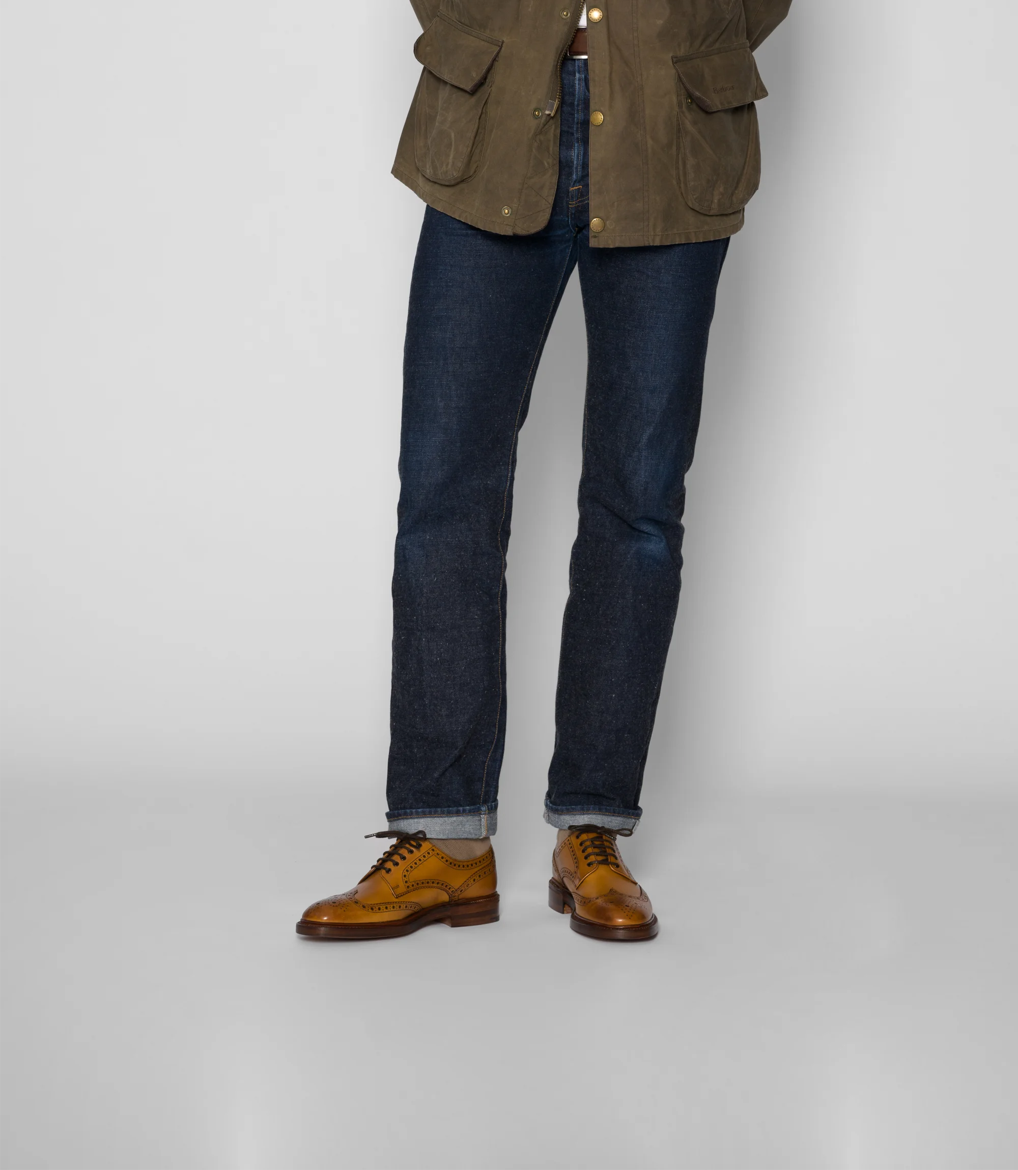 A man wearing navy denim jeans standing in a studio with a grey background wearing his Loake Chester Brogues in Tan Calf Leather.