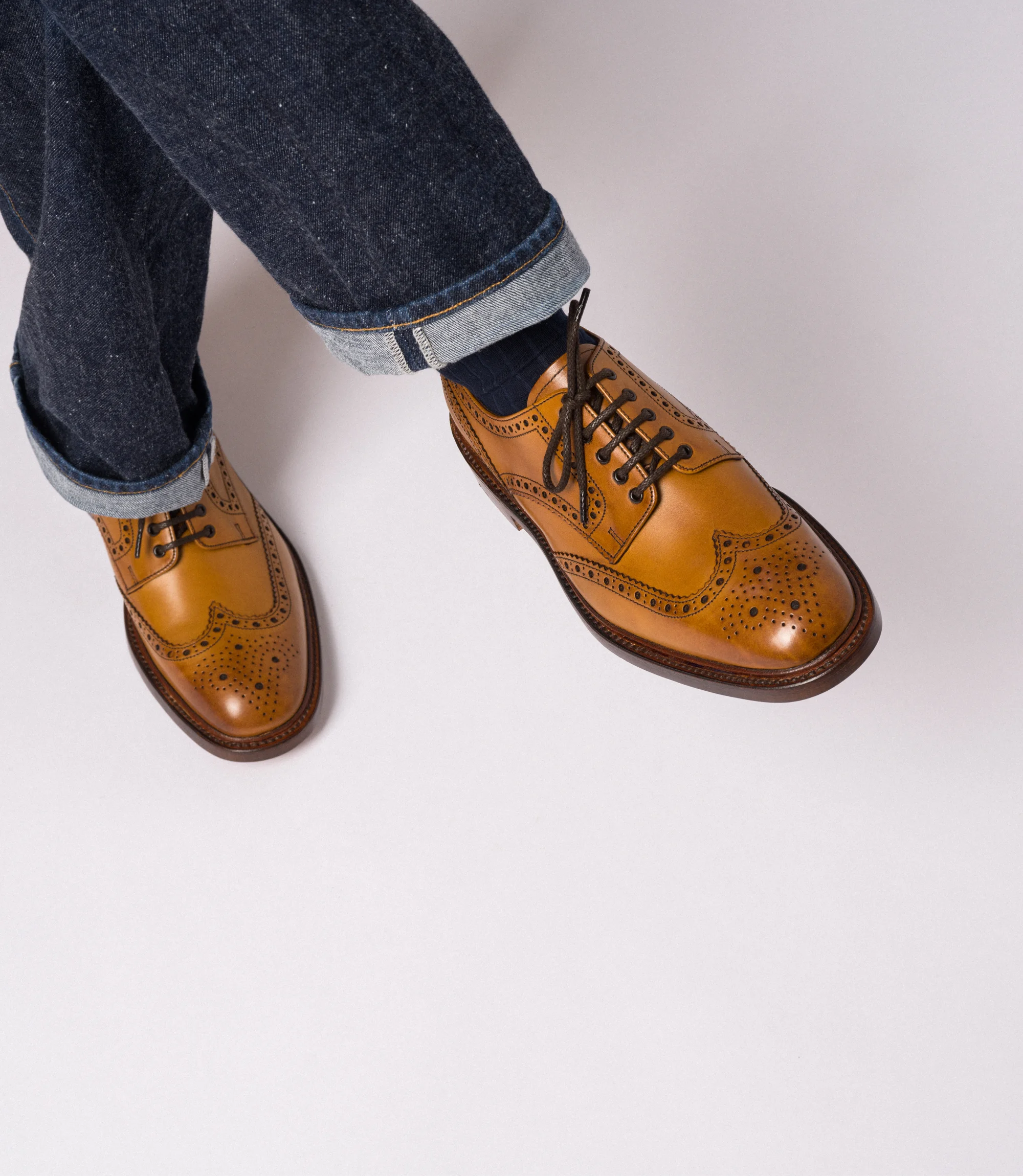 A man wearing navy denim jeans sitting on a chair with legs crossed in a studio with a grey background wearing his Loake Chester Brogues in Tan Calf Leather.