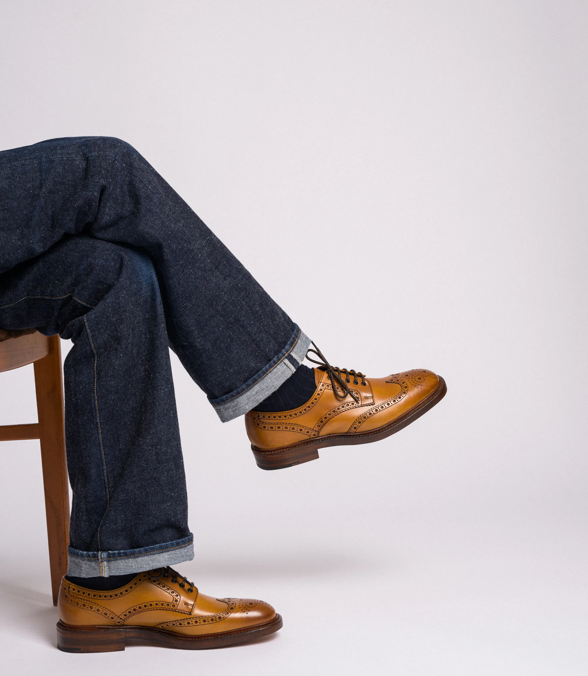 A man wearing navy denim jeans sitting on a chair with legs crossed in a studio with a grey background wearing his Loake Chester Brogues in Tan Calf Leather.