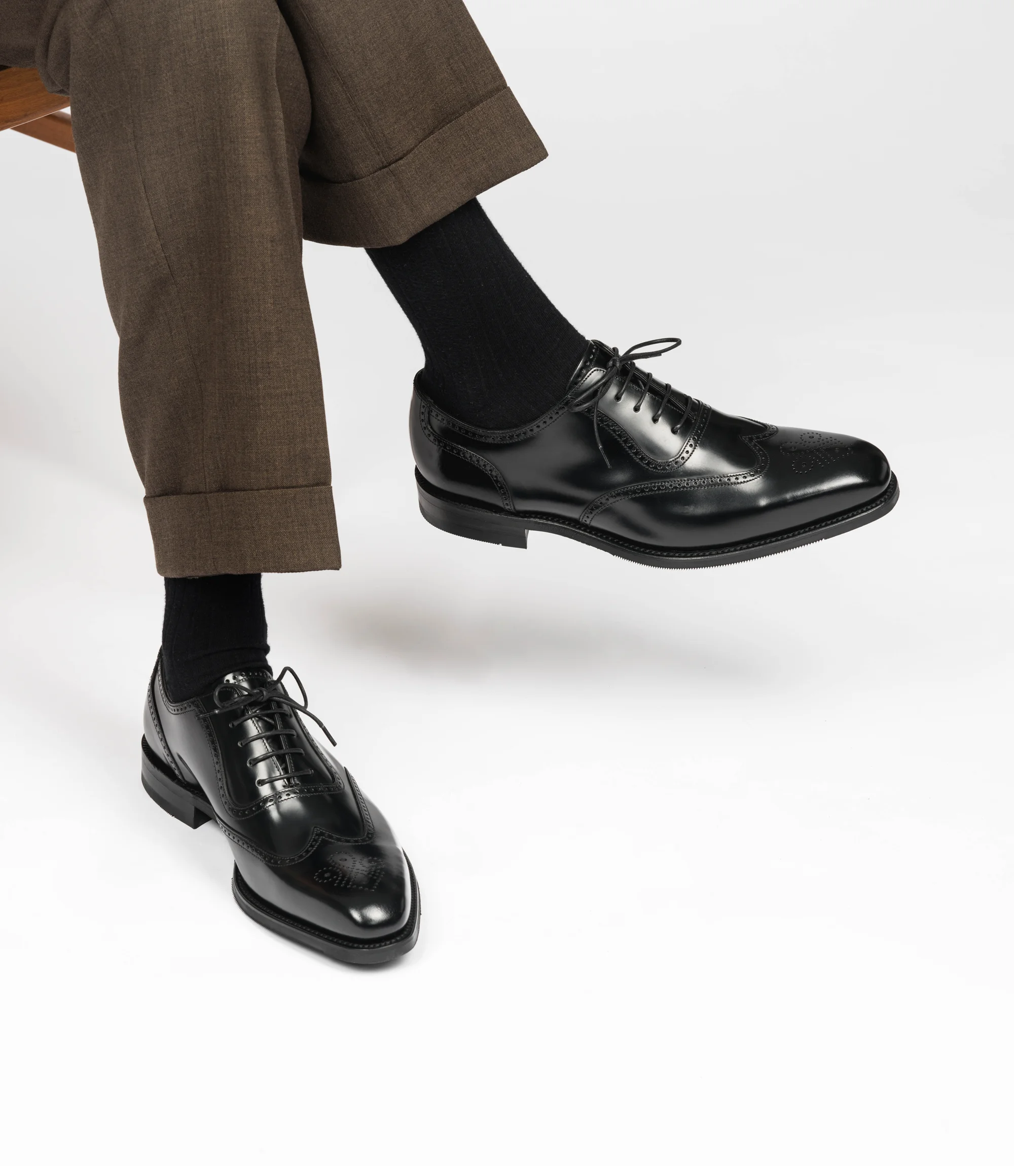 A man wearing brown trousers sitting on a chair with legs crossed in a studio with a grey background wearing his Loake Tay Brogues in Black Calf Leather.