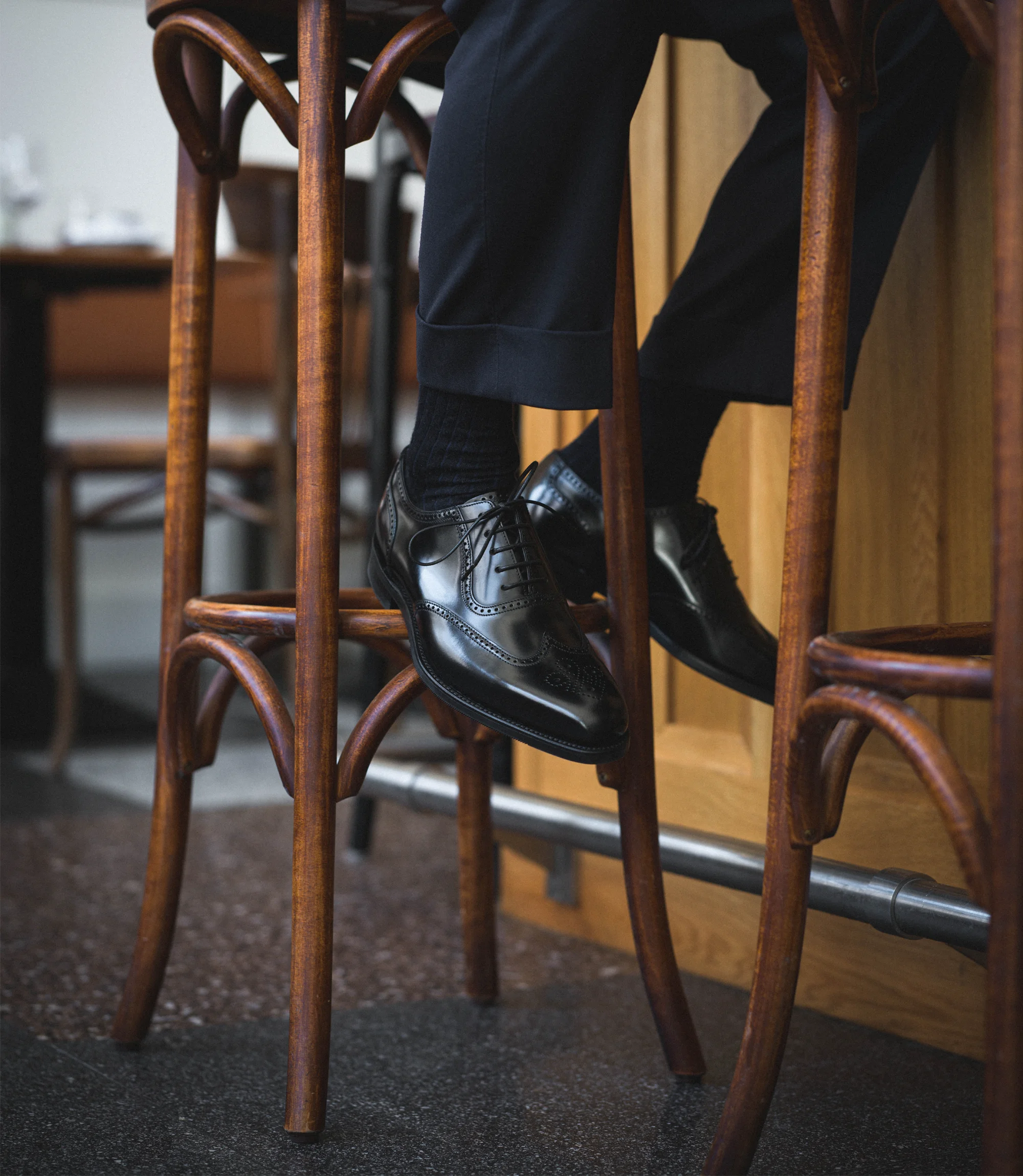 A man wearing navy trousers sitting on a stool wearing his Loake Tay Brogues in Black Calf Leather.