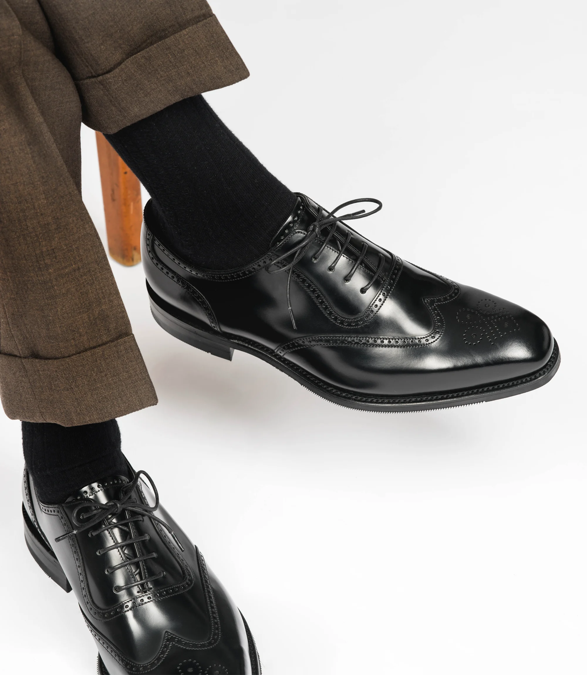 A man wearing brown trousers sitting on a chair with legs crossed in a studio with a grey background wearing his Loake Tay Brogues in Black Calf Leather.
