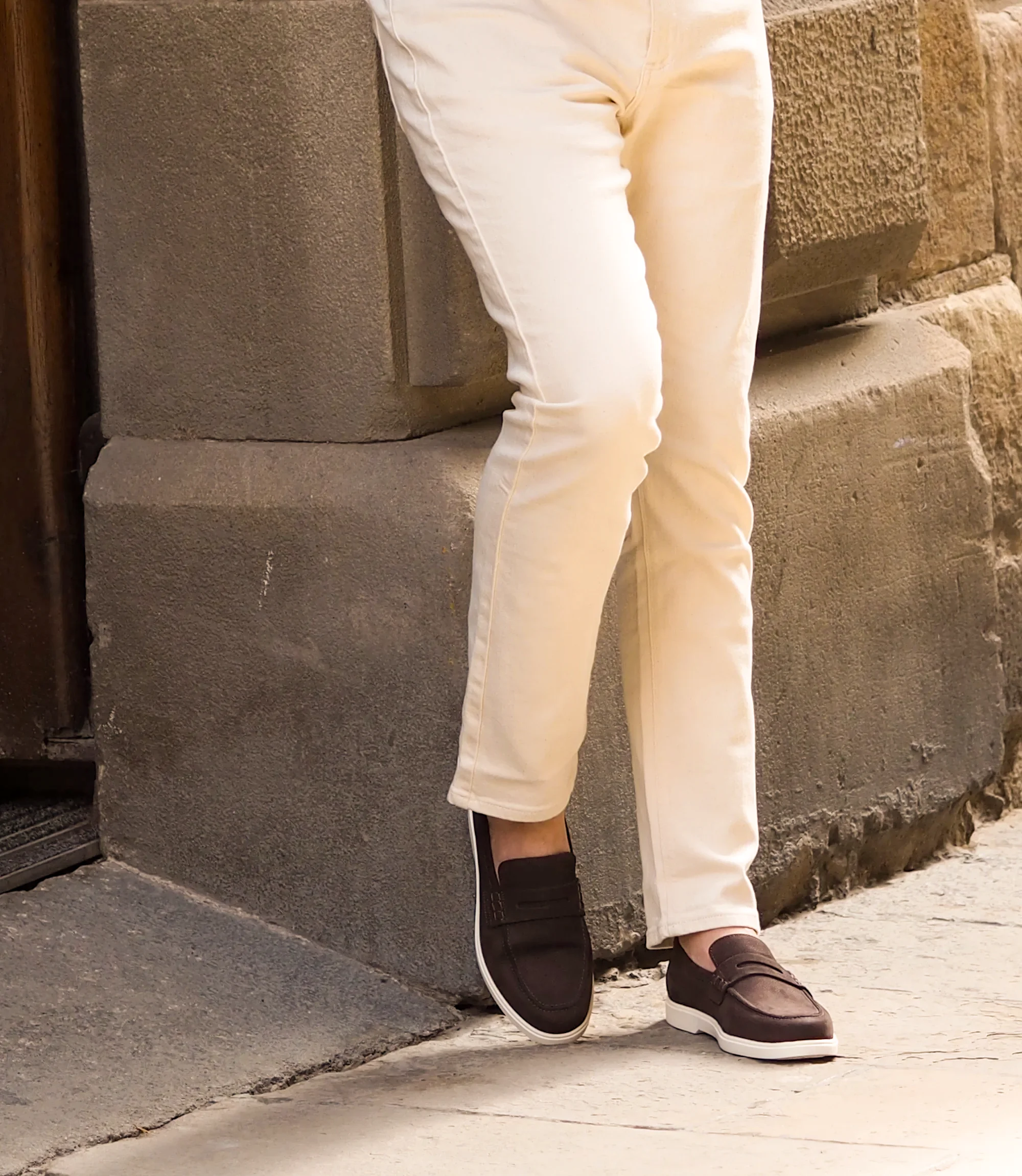 A man wearing white denim trousers standing against a brick wall, wearing Loake Como Loafers in Dark Brown.