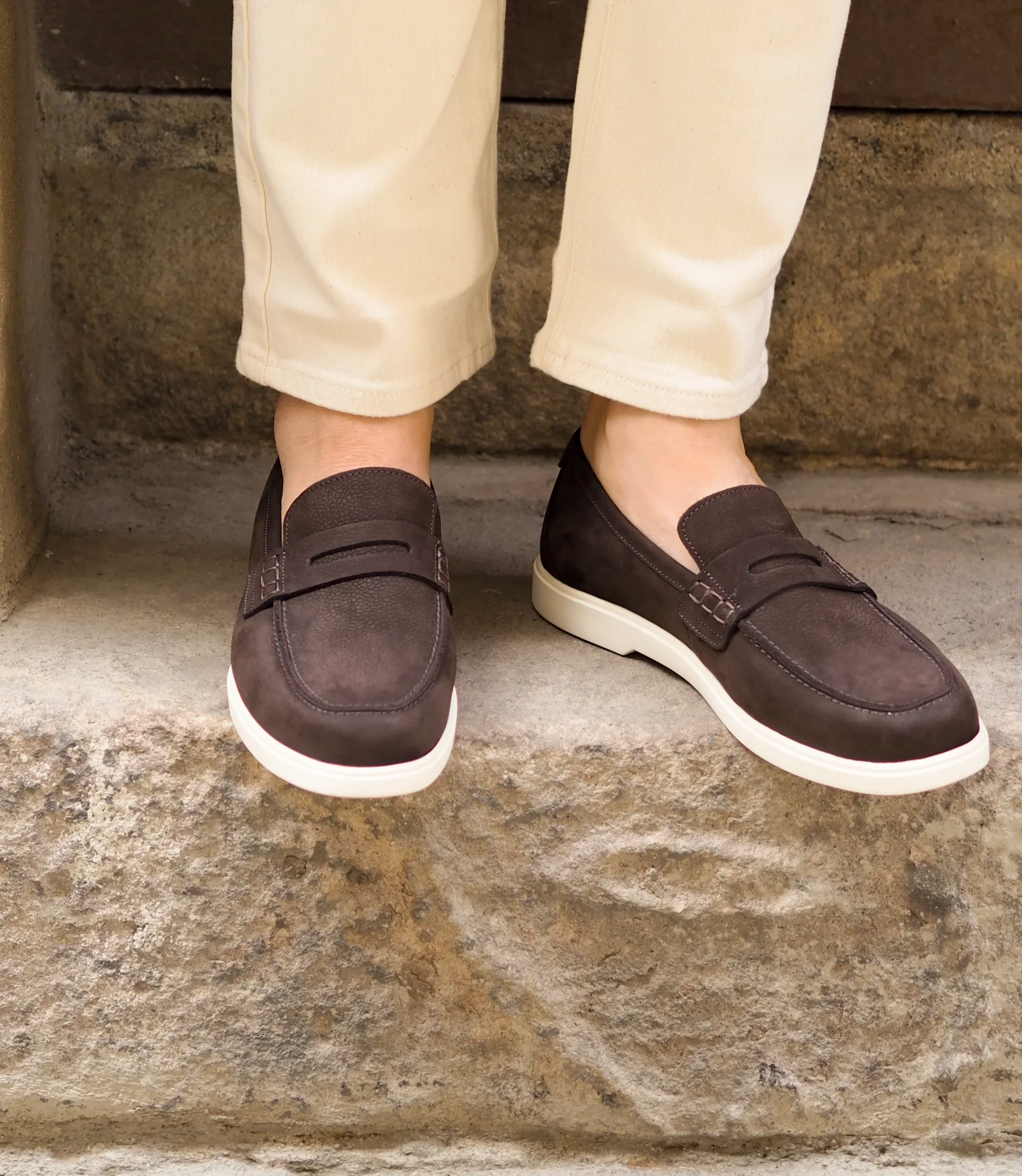A man wearing white denim trousers standing on a concrete step, wearing Loake Como Loafers in Dark Brown.