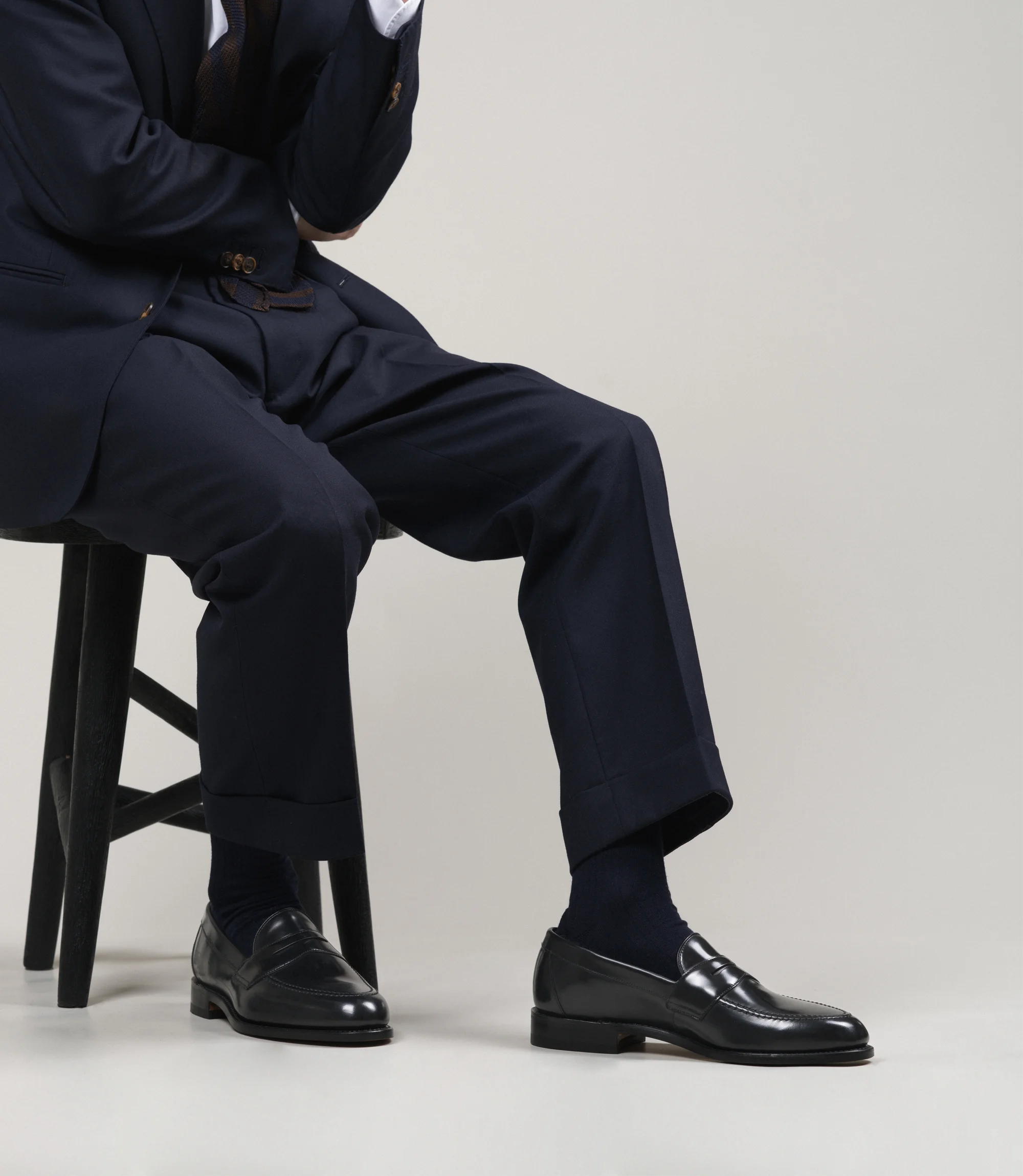 A man wearing navy trousers sitting on a chair in a grey studio backdrop wearing his Loake Imperial Loafers in Black Leather.