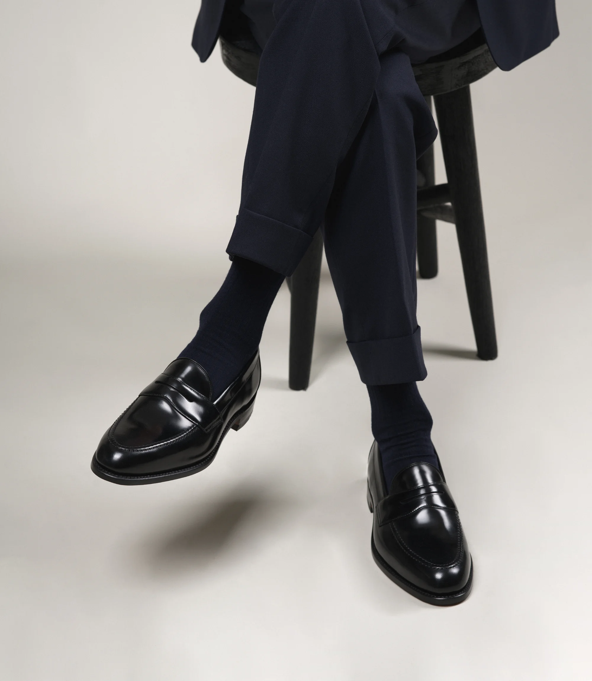 A man wearing navy trousers sitting on a chair in a grey studio backdrop wearing his Loake Imperial Loafers in Black Leather.