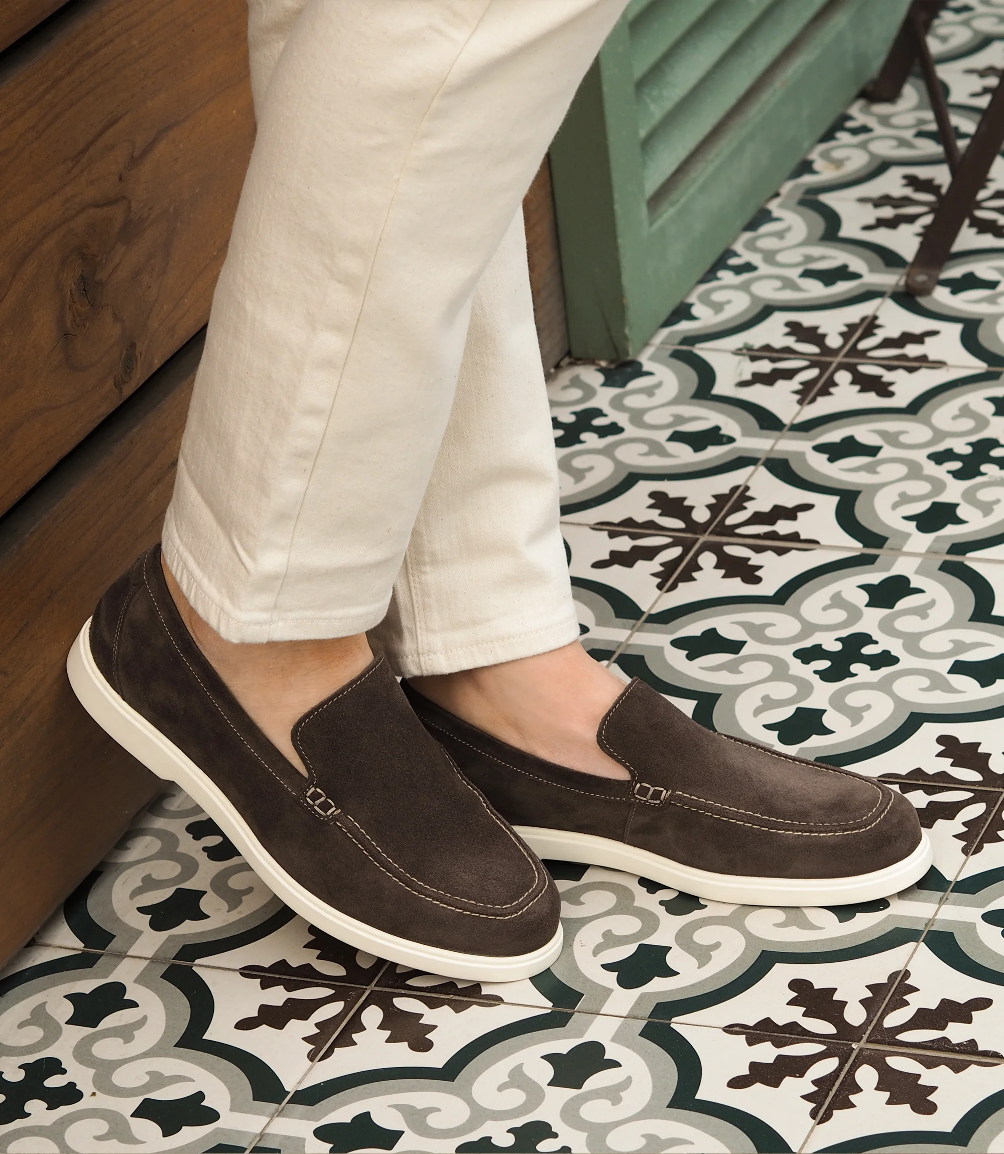A man wearing white denim trousers standing against a wooden wall wearing Loake Tuscany Loafers in Dark Brown Suede.
