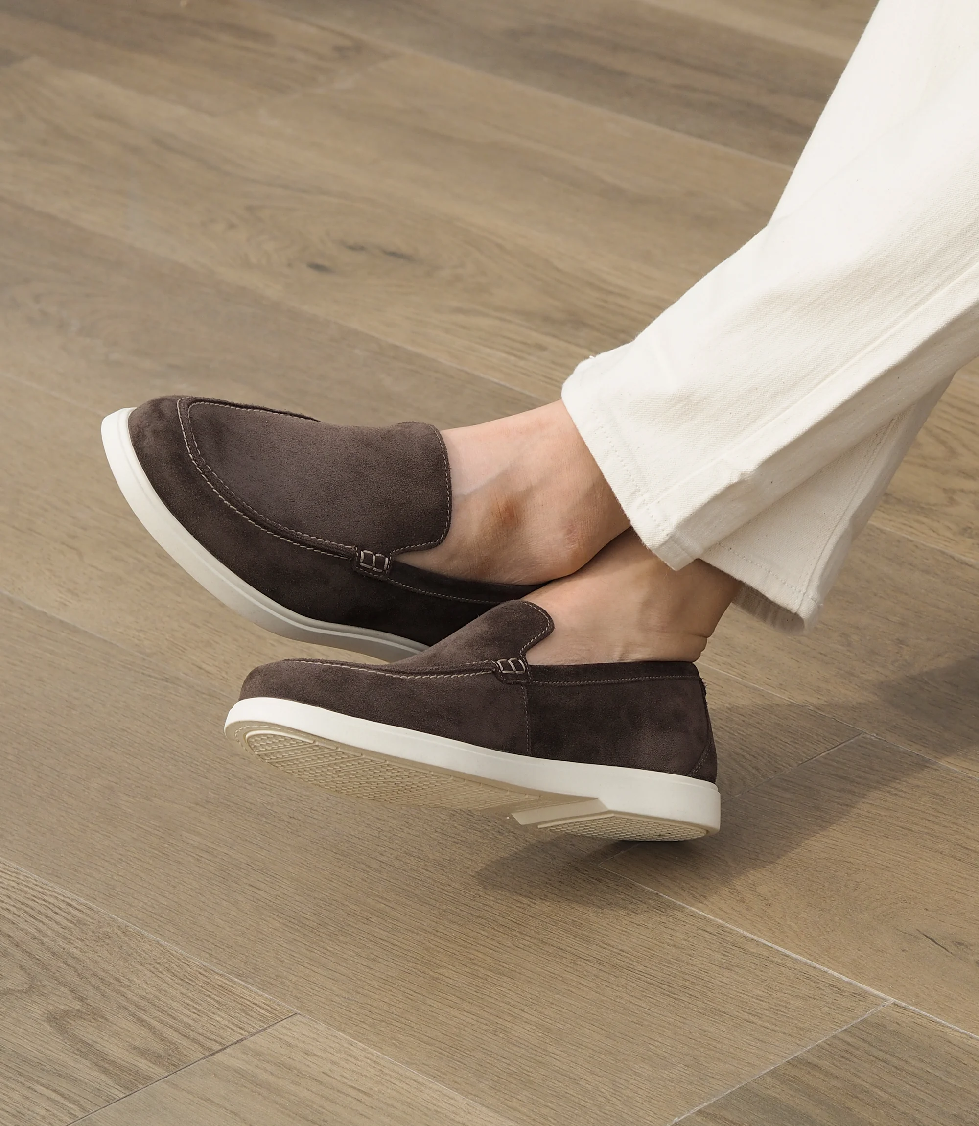 A man wearing white denim jeans sitting on a black sofa in a living room, whilst wearing his Loake Tuscany Loafers in Dark Brown Suede.