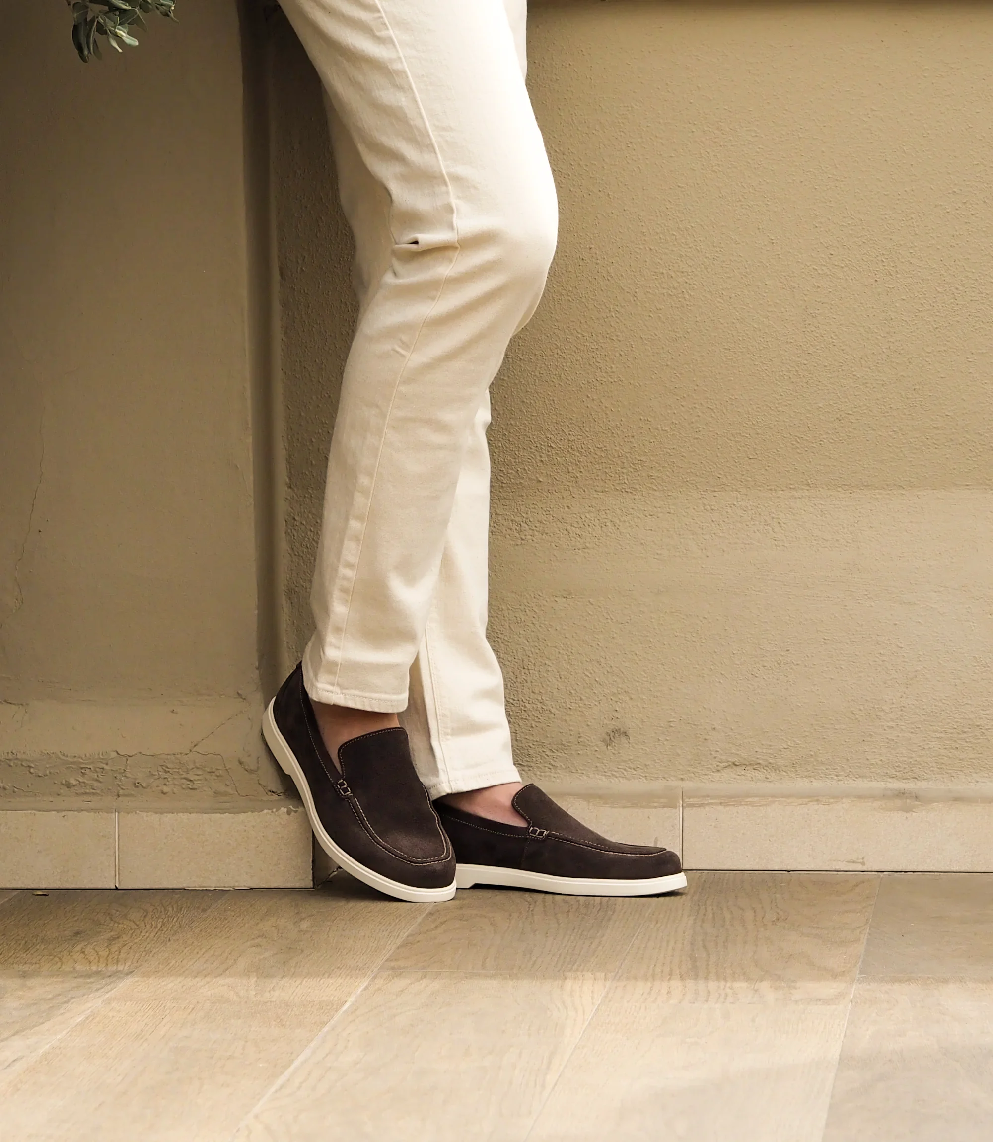 A man wearing white denim trousers standing against a cream brick wall, wearing Loake Tuscany Loafers in Dark Brown.