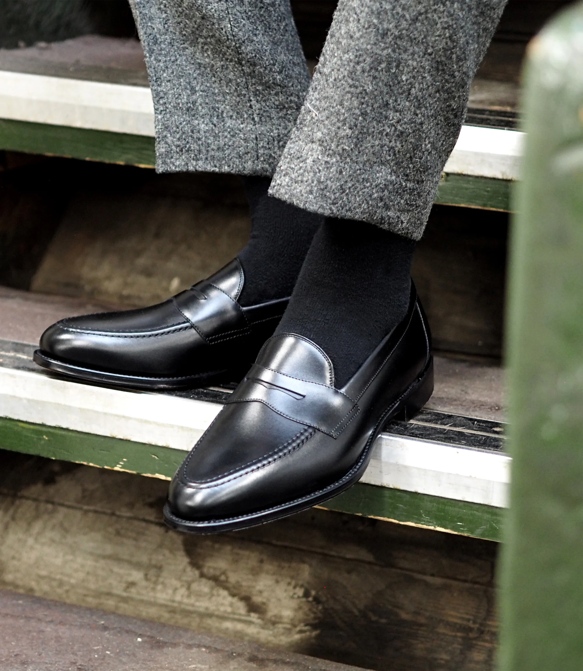 A man wearing grey work trousers sitting on wooden stairs, wearing Loake Hornbeam Loafers in Black leather.
