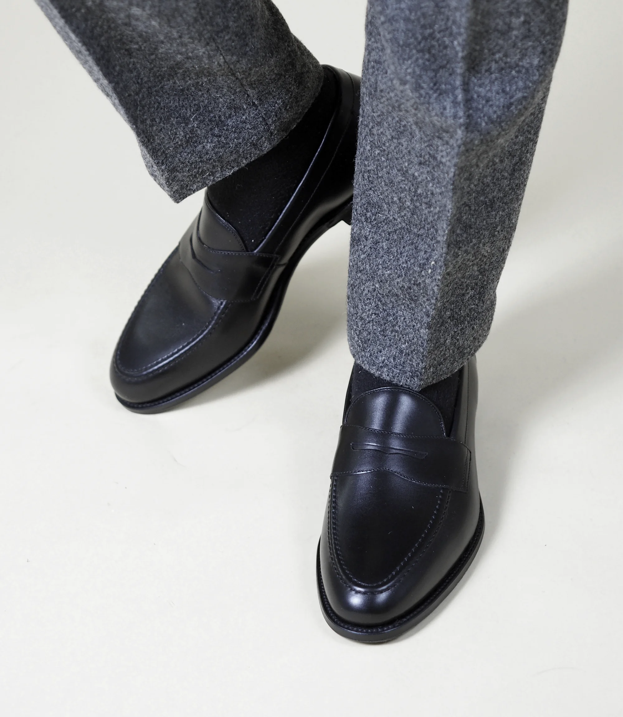 A man wearing grey work trousers sitting standing on a studio floor, wearing Loake Hornbeam Loafers in Black leather.