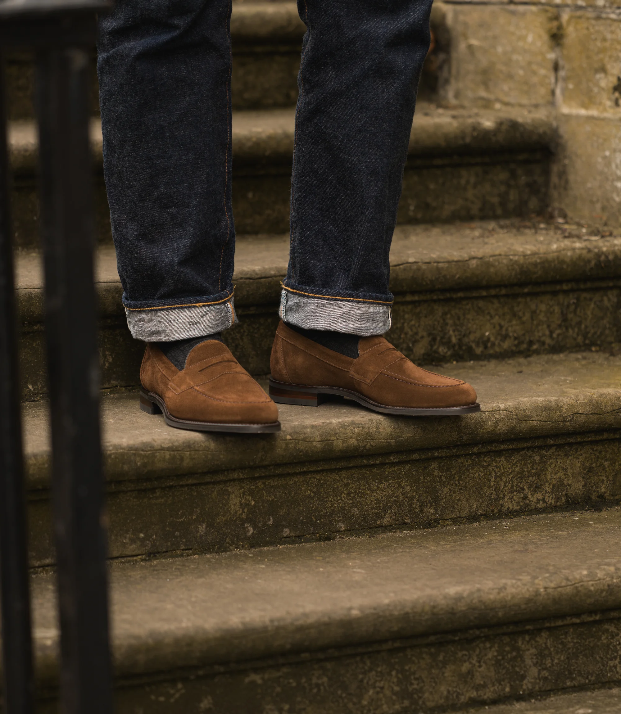 A man wearing blue denim trousers standing on concrete stairs wearing his Loake Imperial Loafers in Brown Suede.
