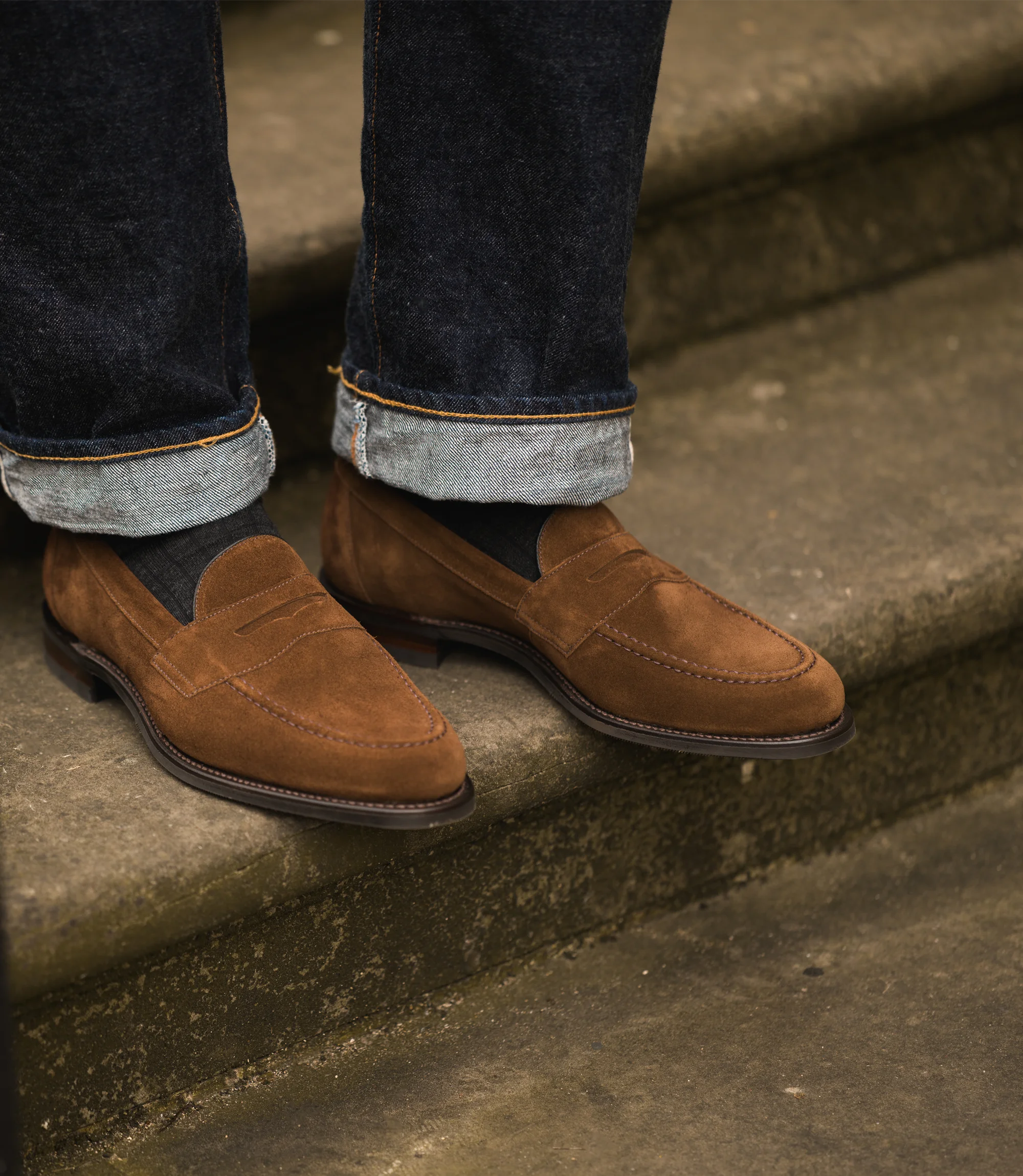 A man wearing blue denim trousers standing on concrete stairs wearing his Loake Imperial Loafers in Brown Suede.