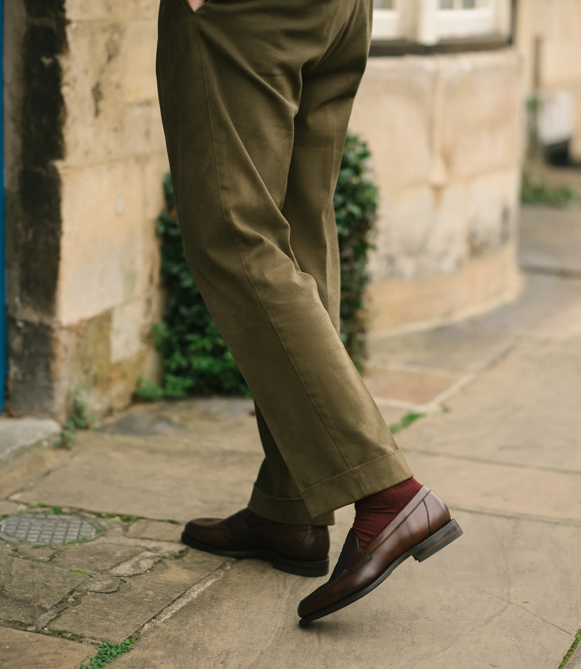A man wearing green trousers walking on concrete floor wearing his Loake Imperial Loafers in Dark brown leather.