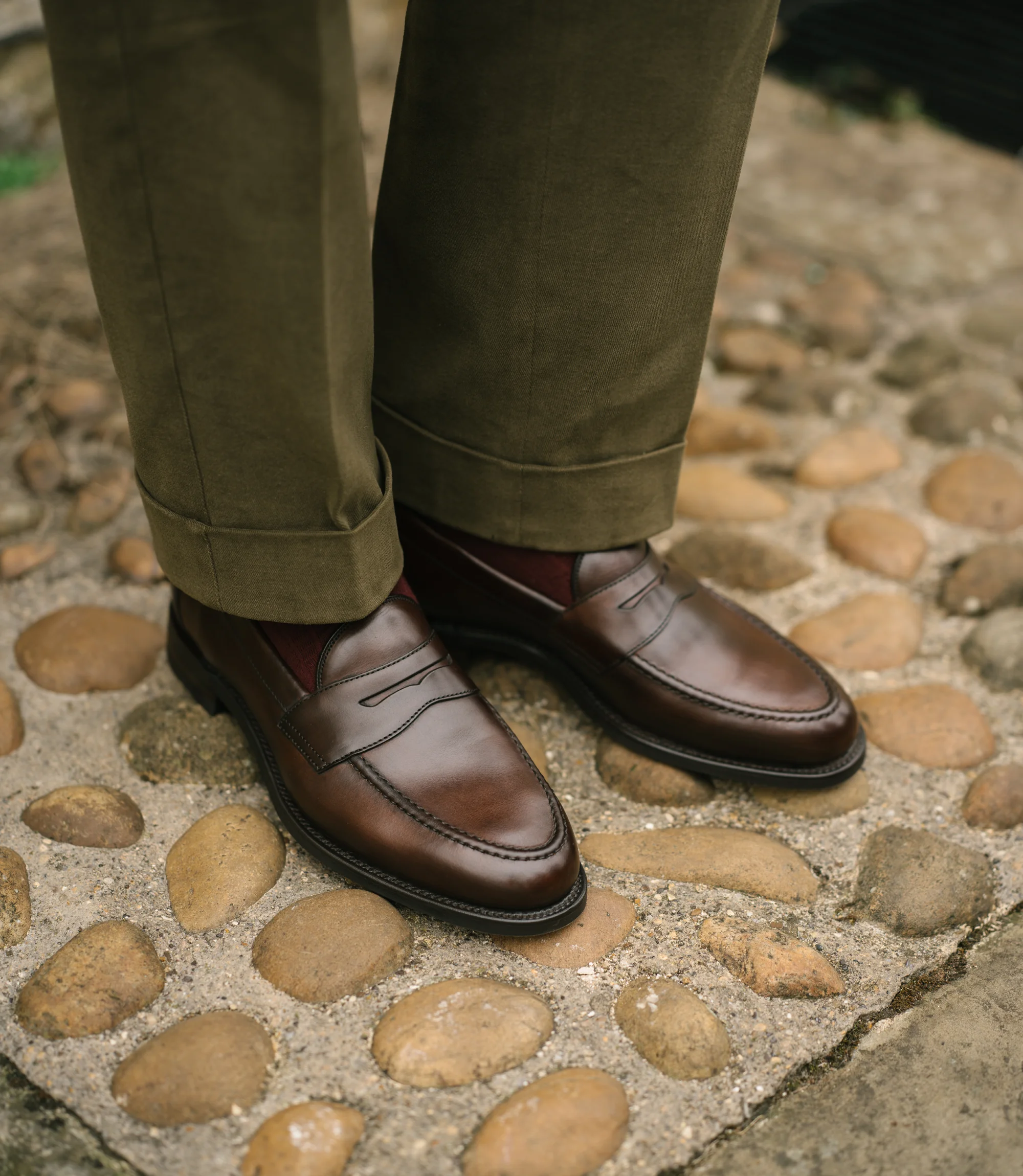 A man wearing green trousers standing on concrete pebbled floor wearing his Loake Imperial Loafers in Dark brown leather.