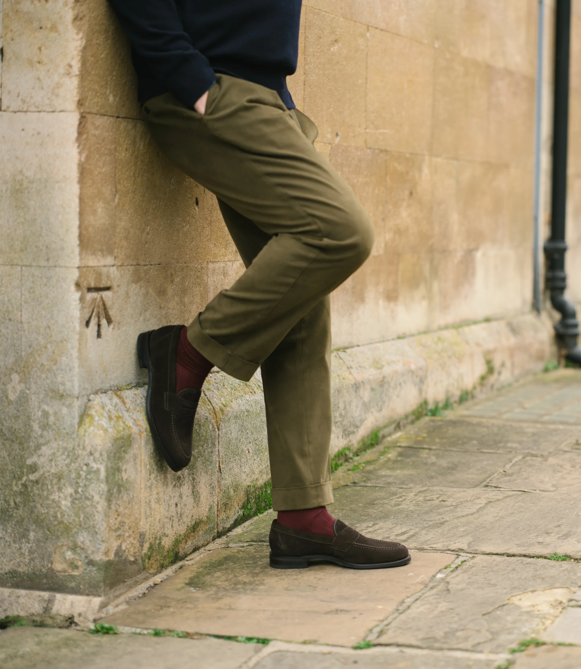 A man leaning up against a brick wall wearing a navy jumper and green trousers wearing his Loake Imperial Loafers in Dark Brown Suede.