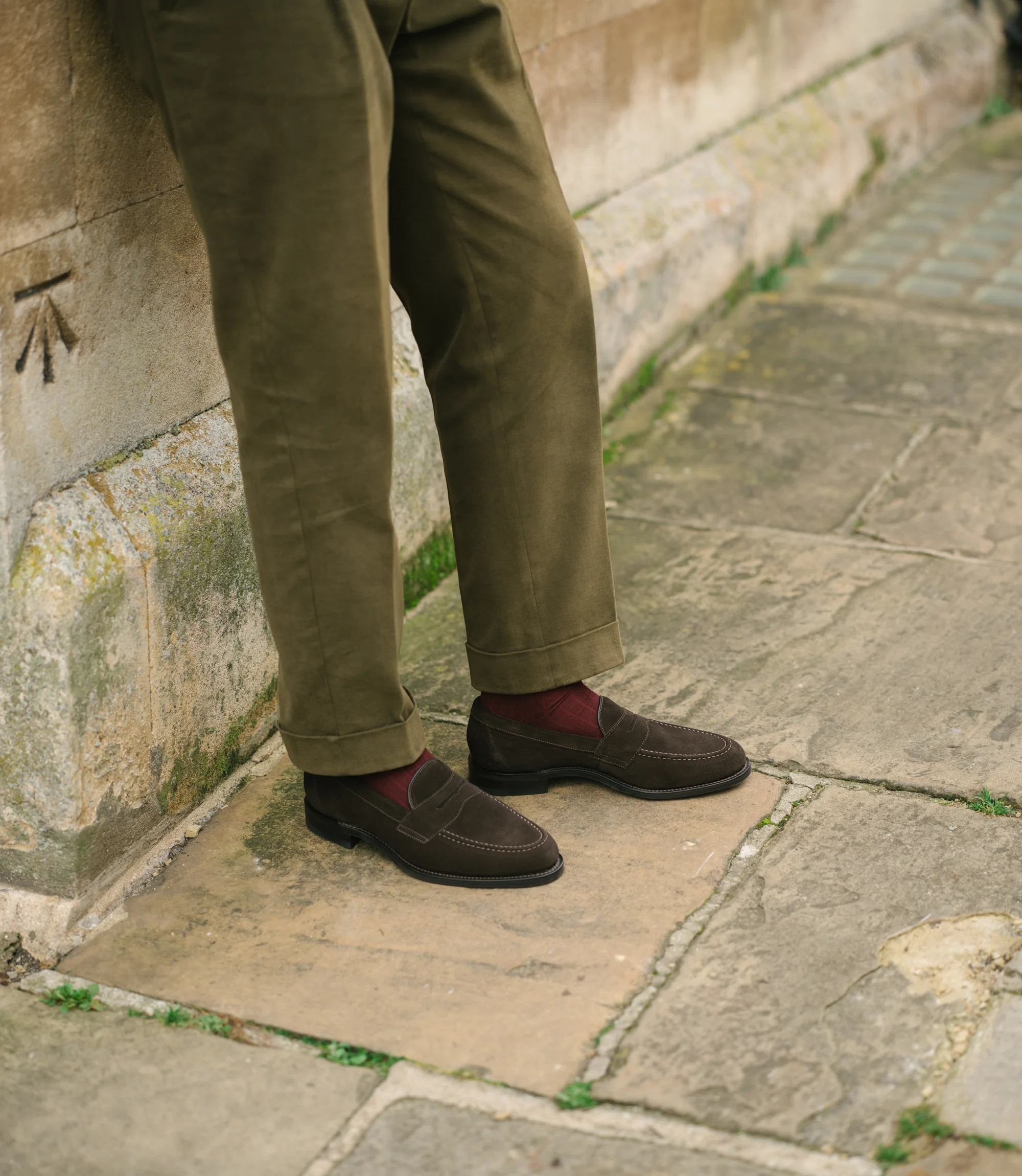 A man leaning up against a brick wall wearing green trousers and wearing his Loake Imperial Loafers in Dark Brown Suede.