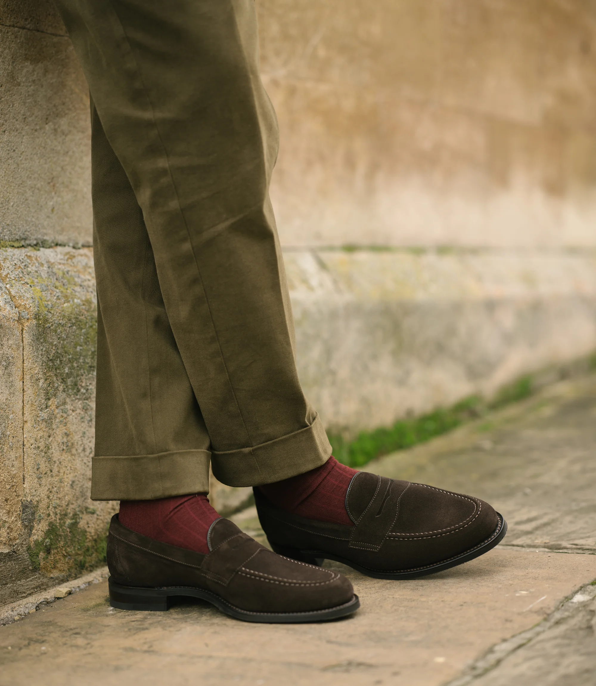 A man leaning up against a brick wall wearing green trousers with his legs crossed is wearing his Loake Imperial Loafers in Dark Brown Suede.