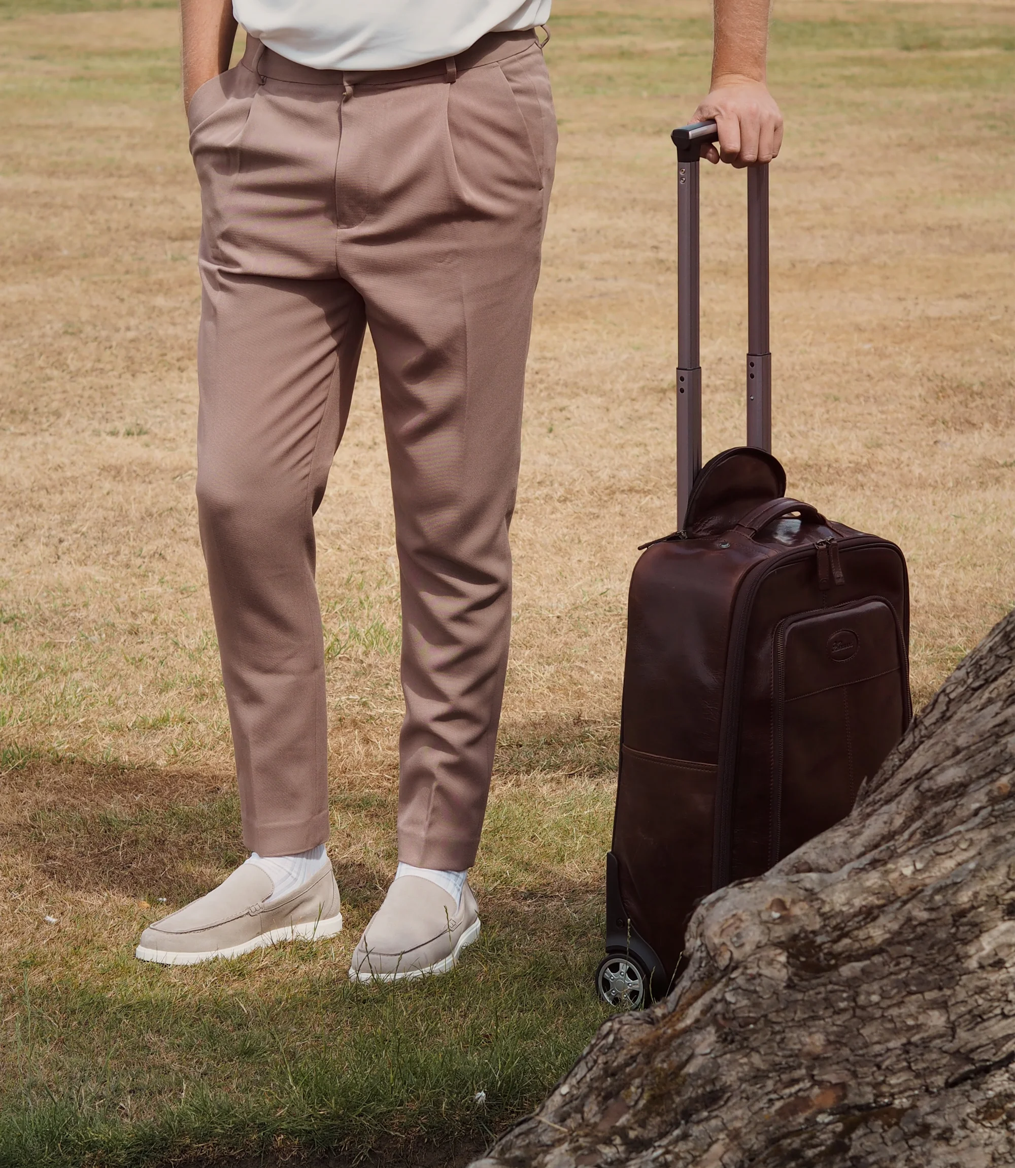A man wearing brown trousers holding a brown travel suitcase standing on grass wearing his Loake Tuscany Loafers in Stone Suede.