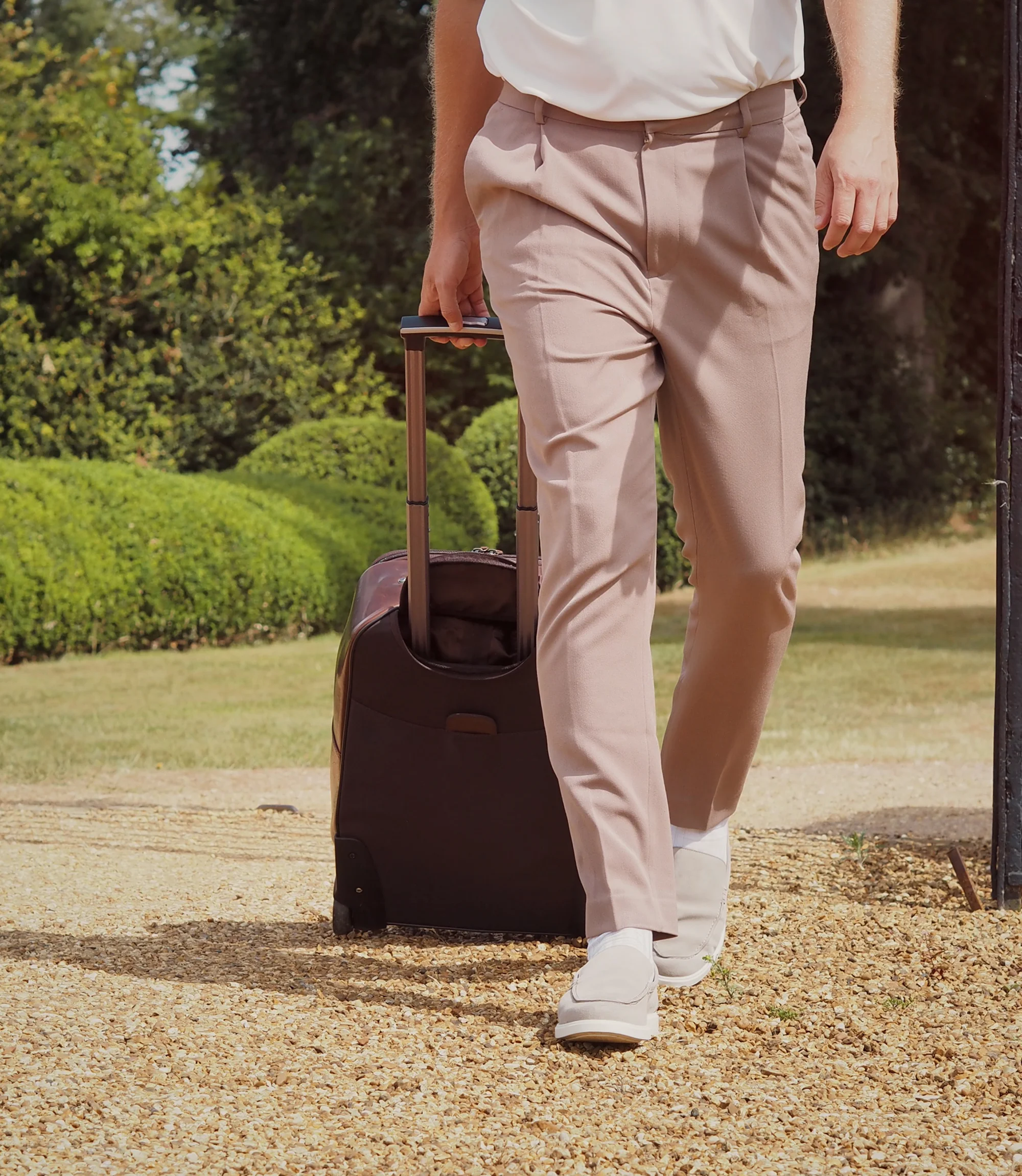A man wearing brown trousers pulling a brown travel suitcase along the pebbles wearing his Loake Tuscany Loafers in Stone Suede.