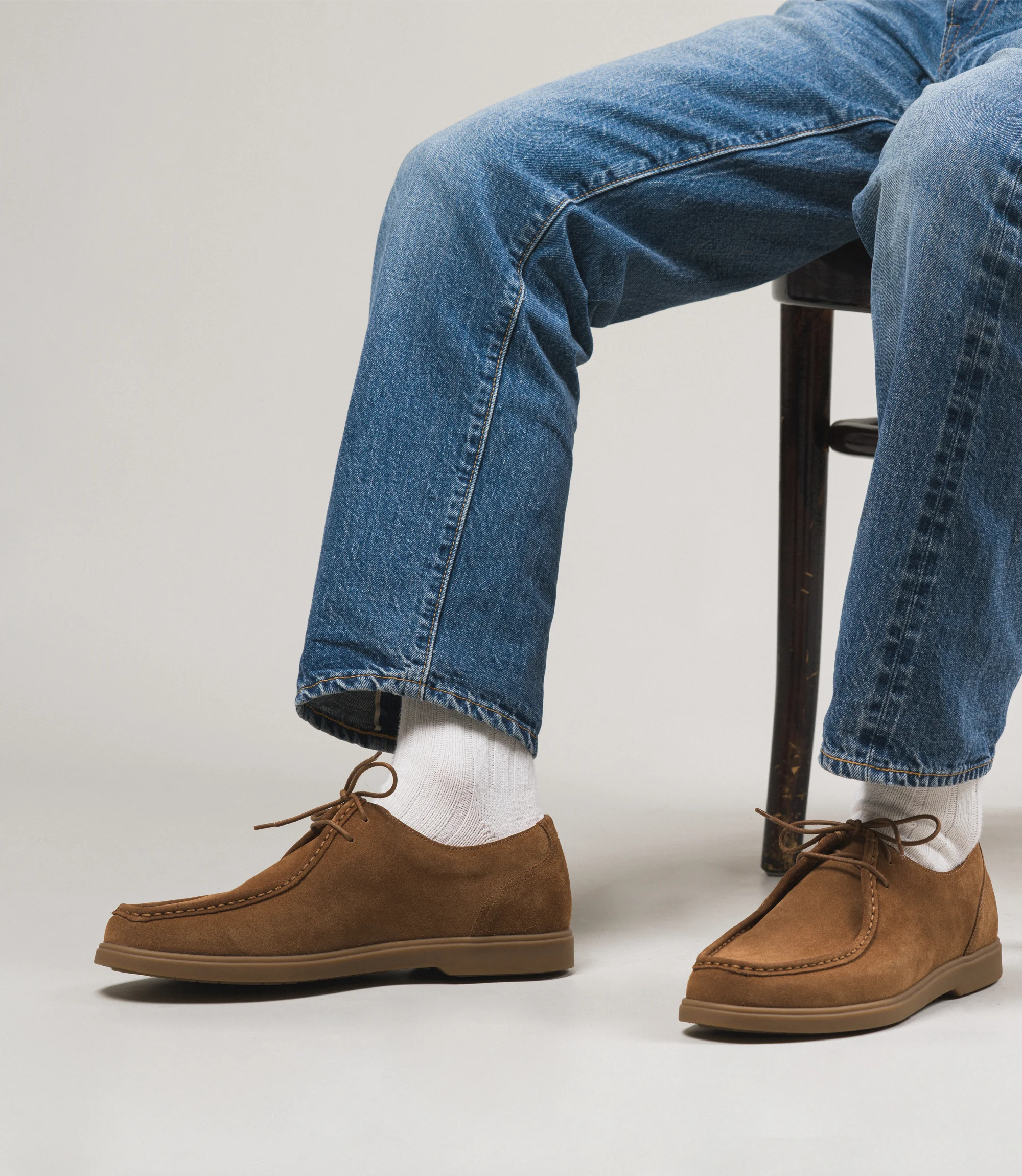 A man wearing light blue denim jeans sat down on chair in a studio with a grey backdrop wearing his Loake Arezzo shoes in Chestnut Suede.
